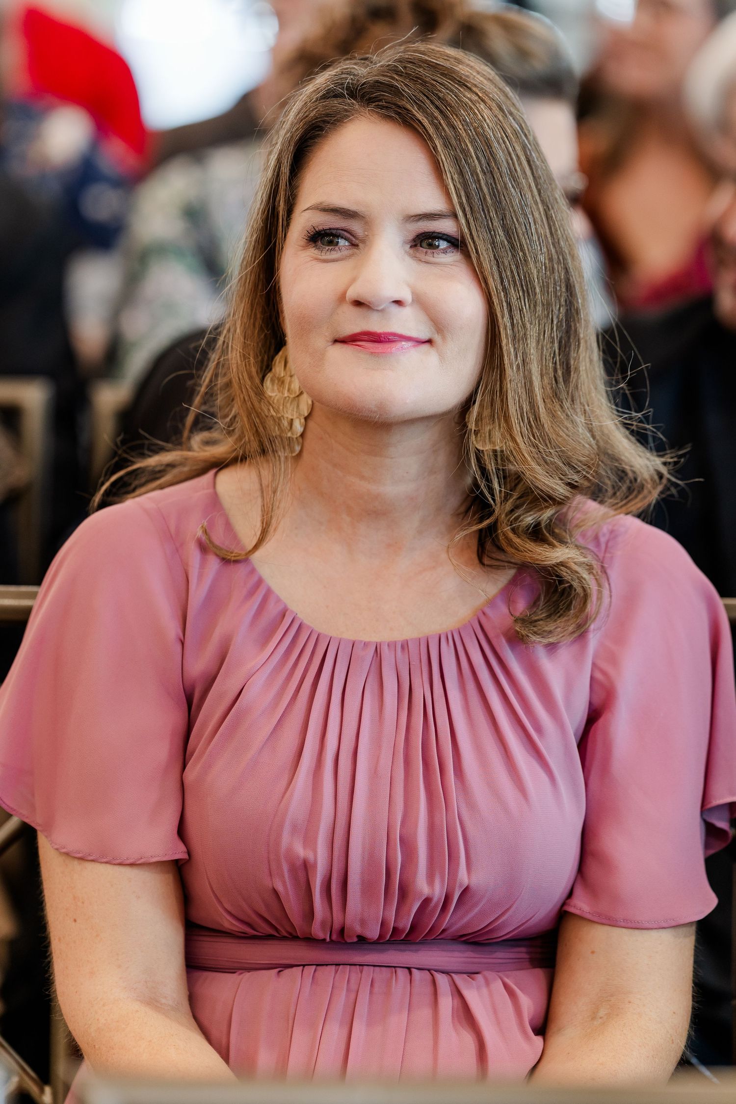 Smiling woman with wavy light brown hair wears gold earrings and a dusty pink flutter-sleeve dress while seated among wedding guests.