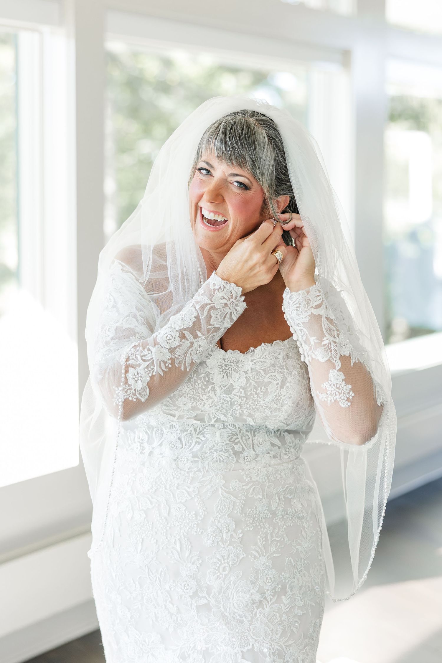 Smiling bride in a long-sleeved white lace wedding gown and sheer veil adjusts her earring in a bright, window-lit room.