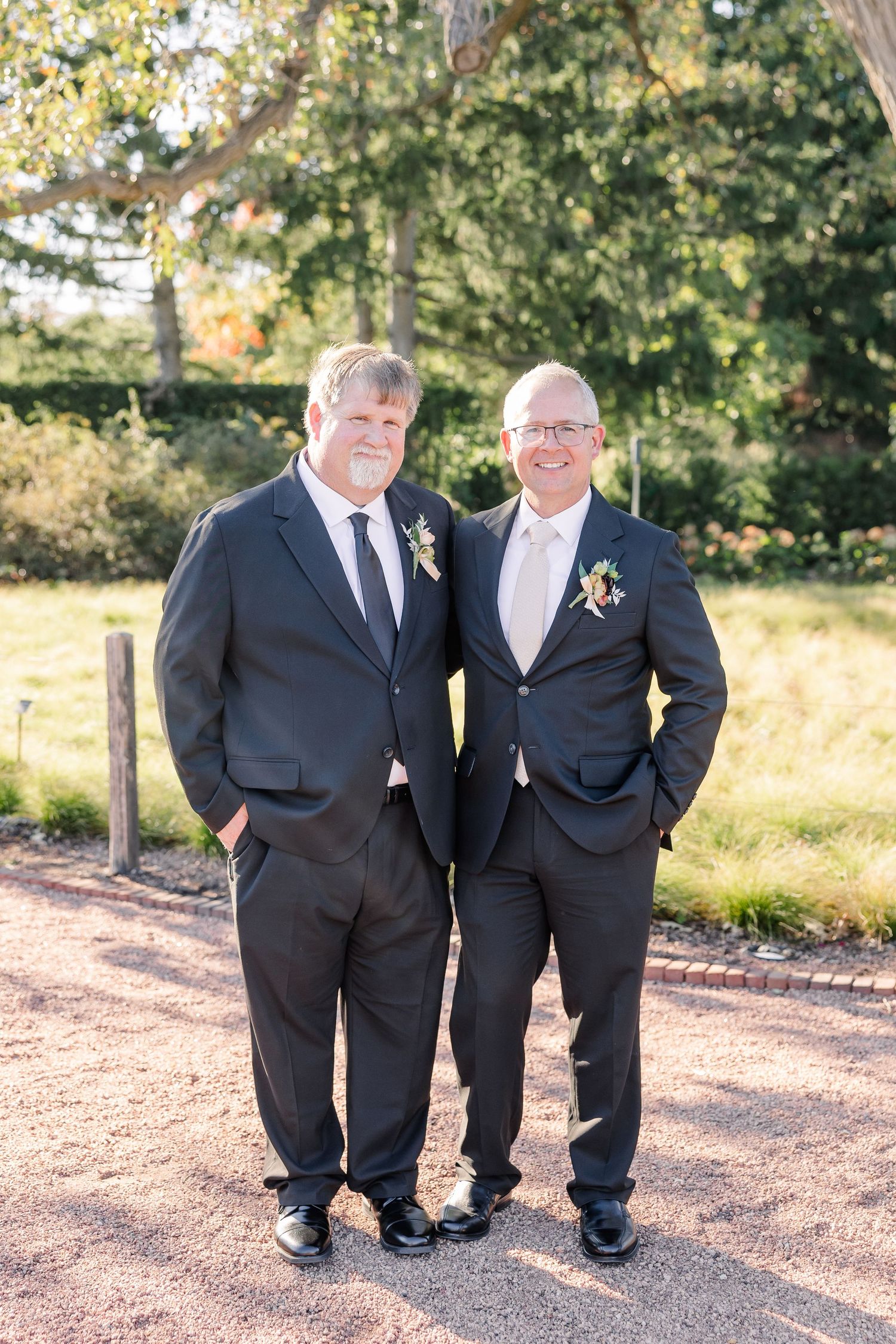 Two men in dark suits with boutonnieres stand side by side smiling on a gravel path in a sunny garden at a wedding.