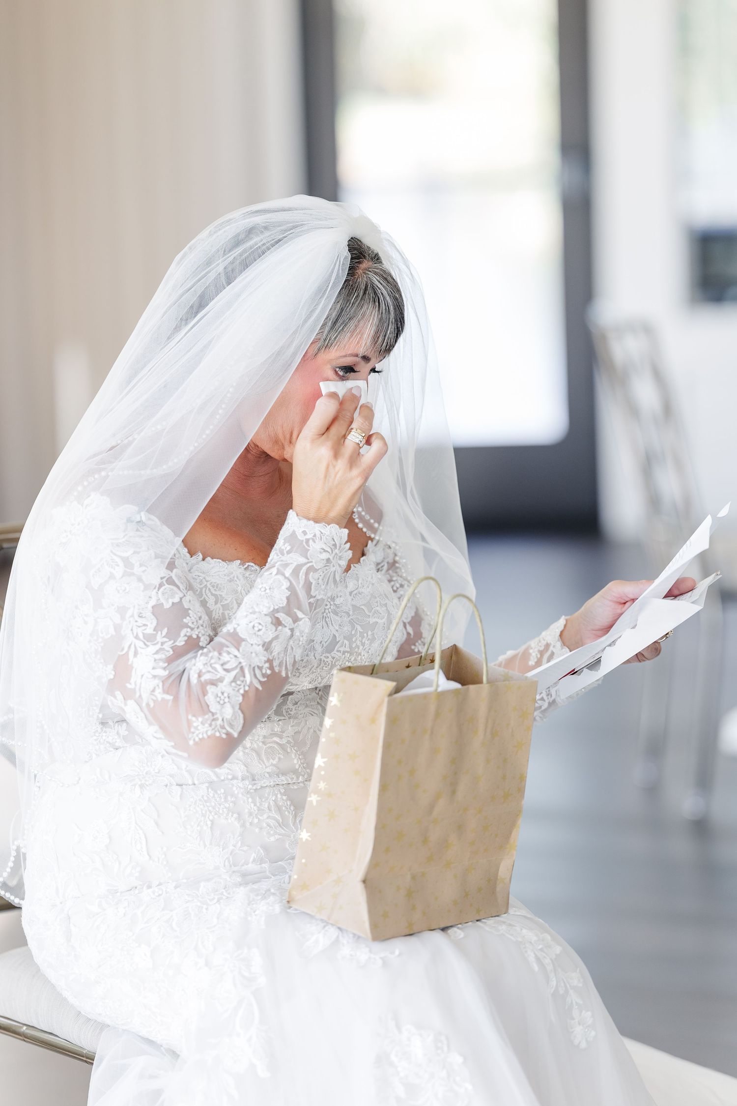 Emotional bride in a lace wedding dress and veil wipes away tears while holding a gift bag and reading a handwritten letter.