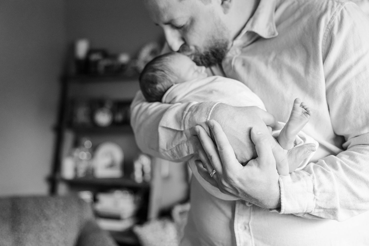 Black and white photo of bearded father gently kissing and cradling newborn baby in arms.