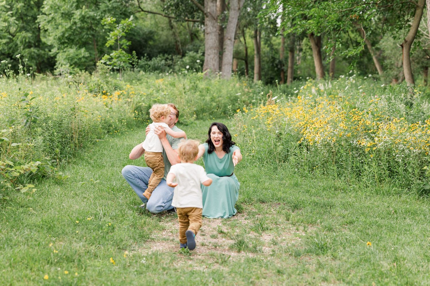 Family playing in a wildflower meadow, mom in green dress laughing as toddler runs toward her.