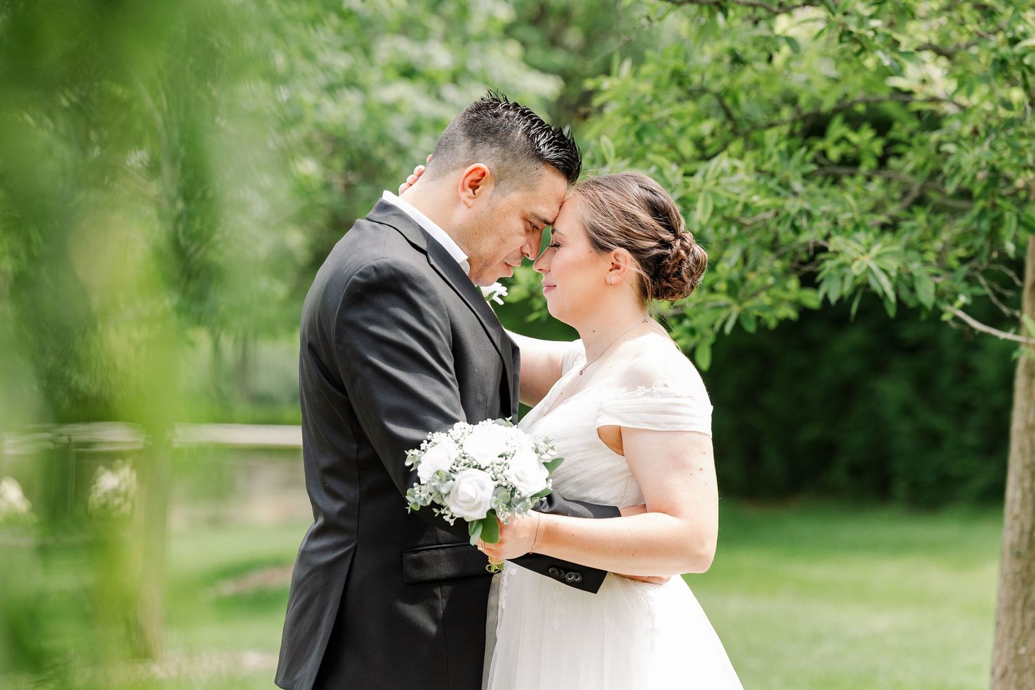 Romantic portrait of Monika and Rafael surrounded by greenery while she holds her white rose bouquet.