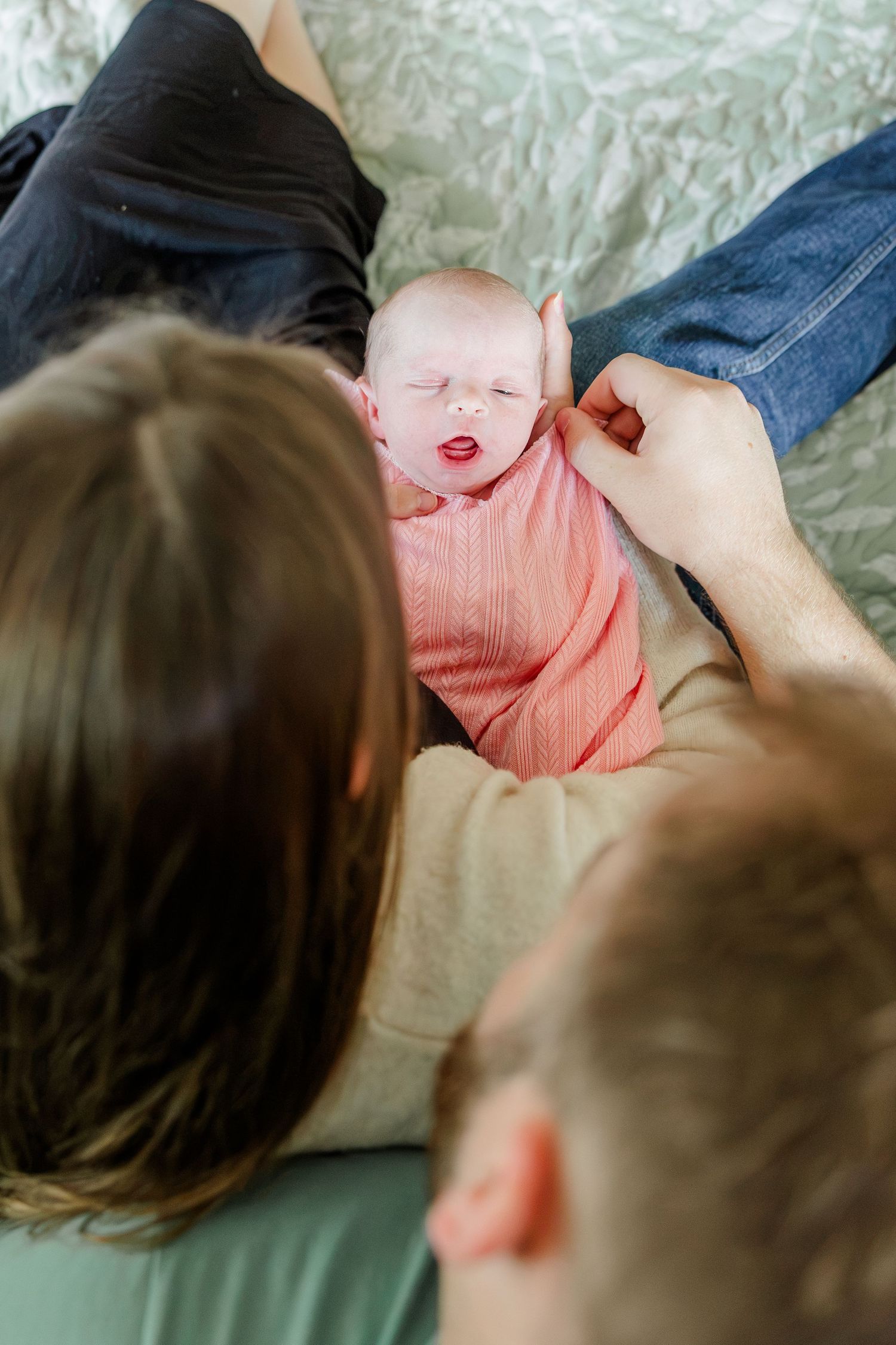 New parents lovingly gaze at their swaddled newborn baby in pink on a bed.