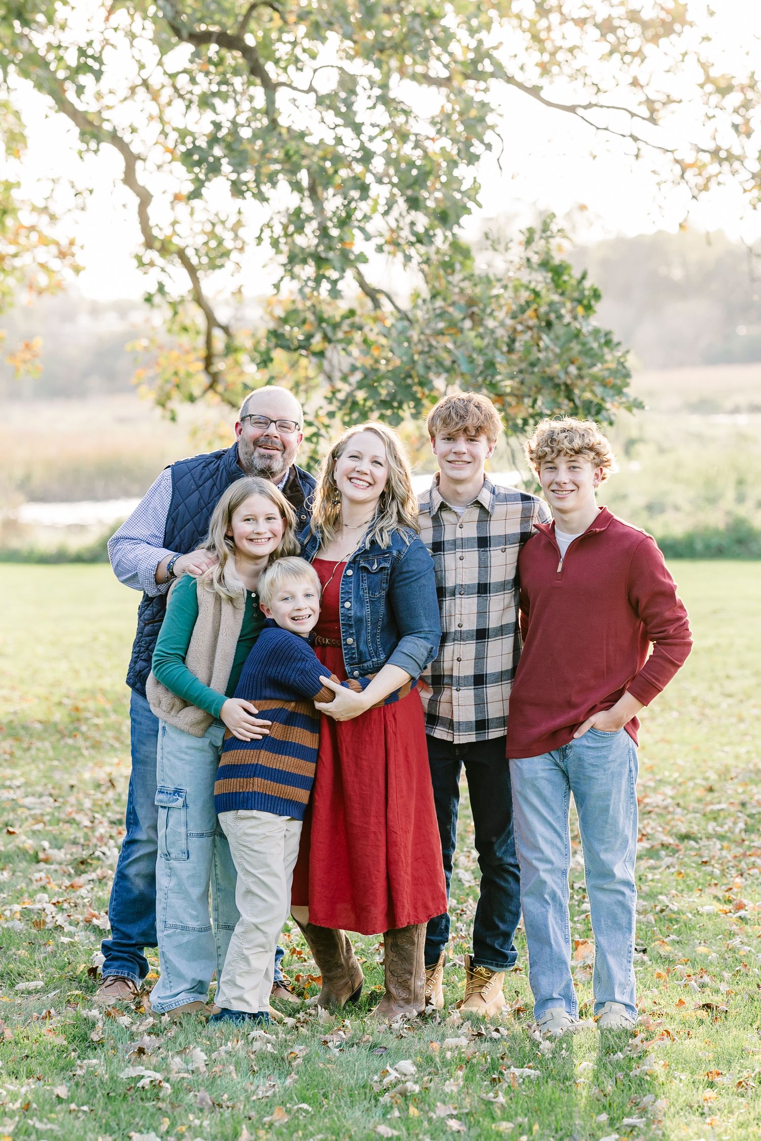 A smiling family of six in fall outfits stands close together on a grassy lawn beneath a leafy tree beside a pond.