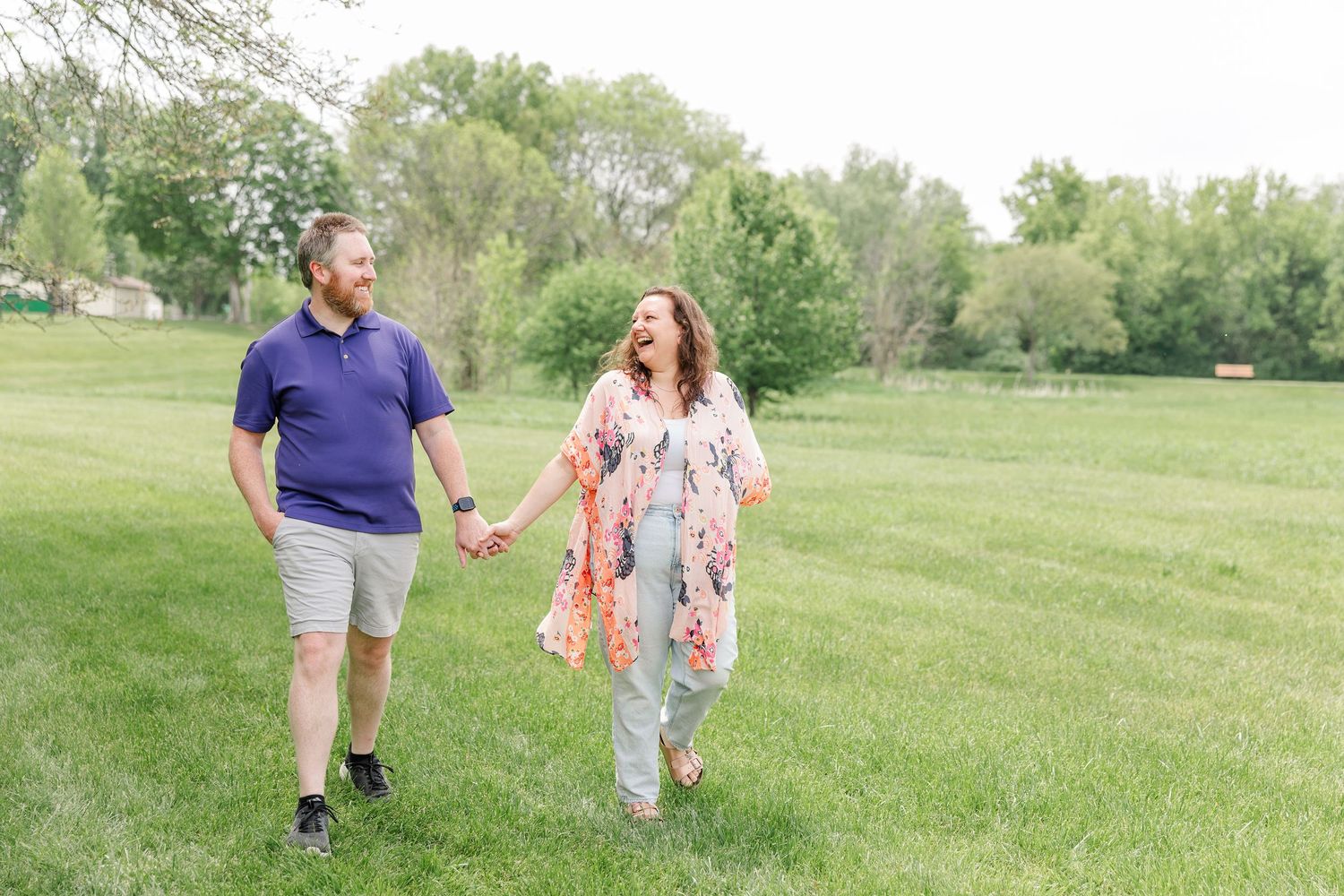 A laughing couple holds hands while walking together across a wide open green field in spring.