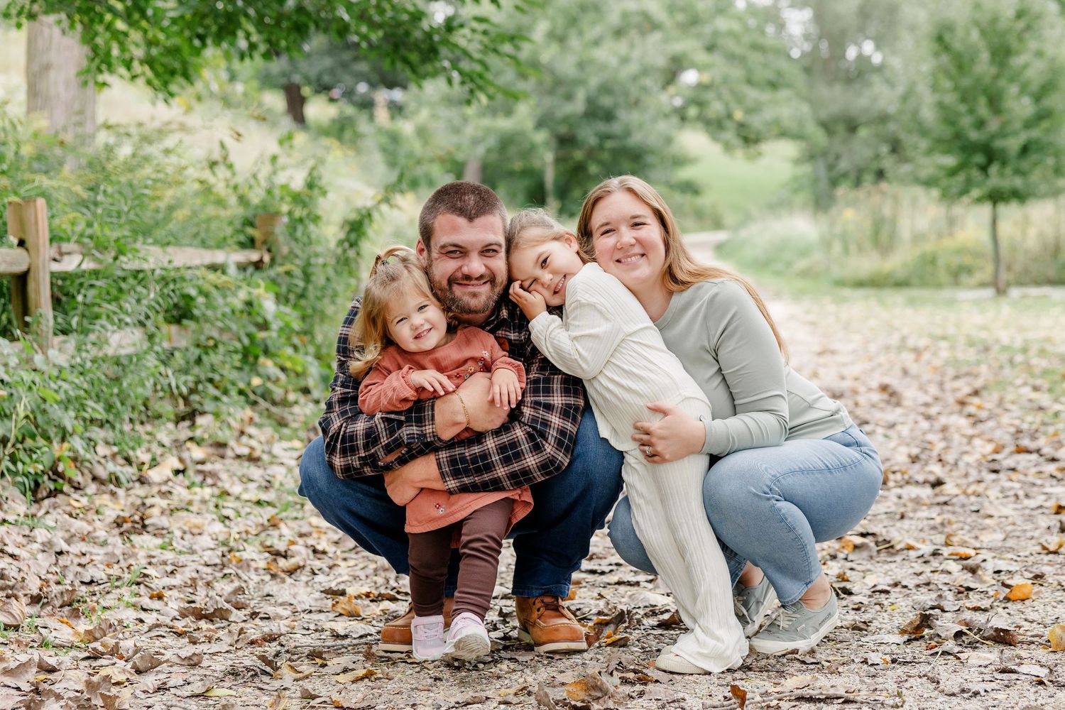 Happy family of four posing outdoors in autumn, parents crouching with two young daughters hugging them among fallen leaves.