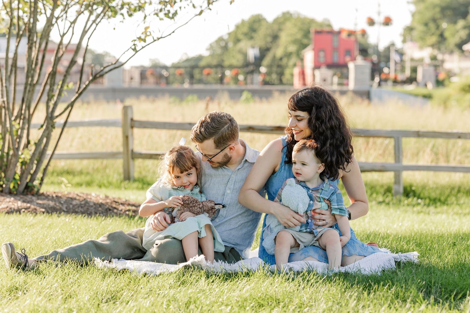 Family of four sitting on grass in a sunny park, parents smiling with two young children on a blanket.