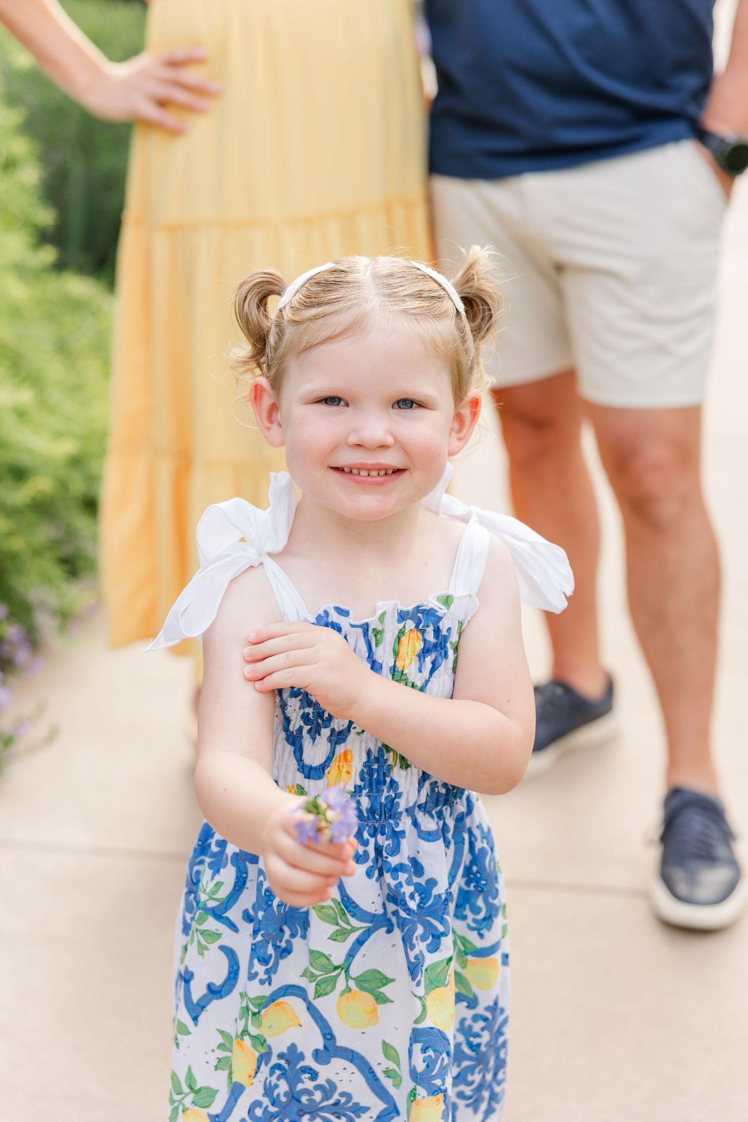 Happy toddler girl in blue floral sundress smiles holding a flower outdoors.