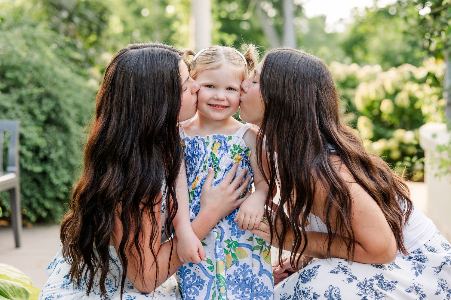 Two dark-haired women kissing smiling toddler girl on cheeks outdoors.