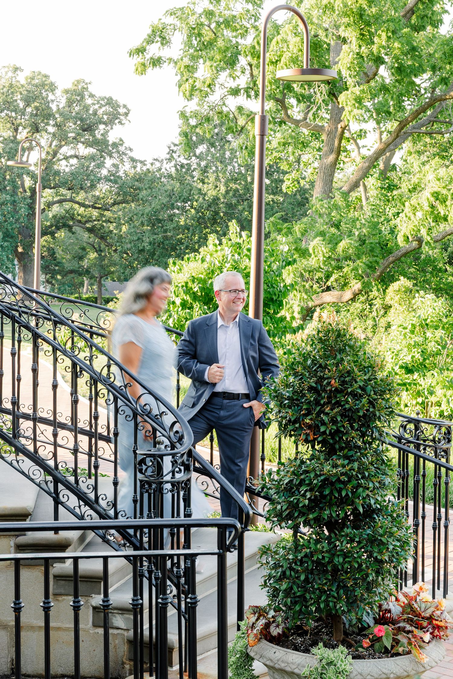 Couple in formal attire descending ornate iron staircase surrounded by lush greenery and decorative lamp post.
