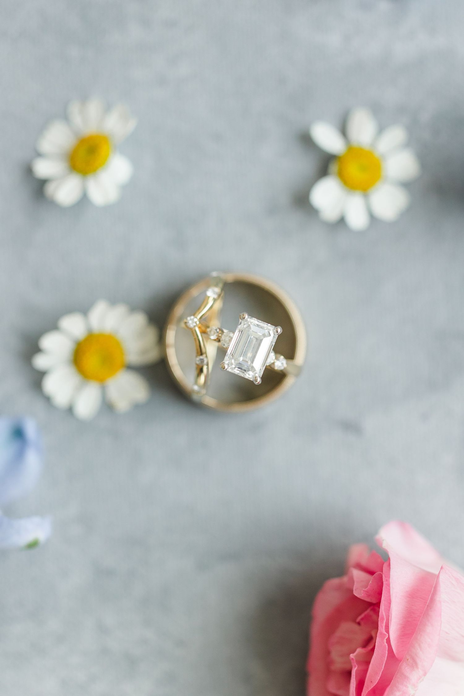 Emerald cut diamond engagement ring in gold box surrounded by white daisy flowers on gray background.