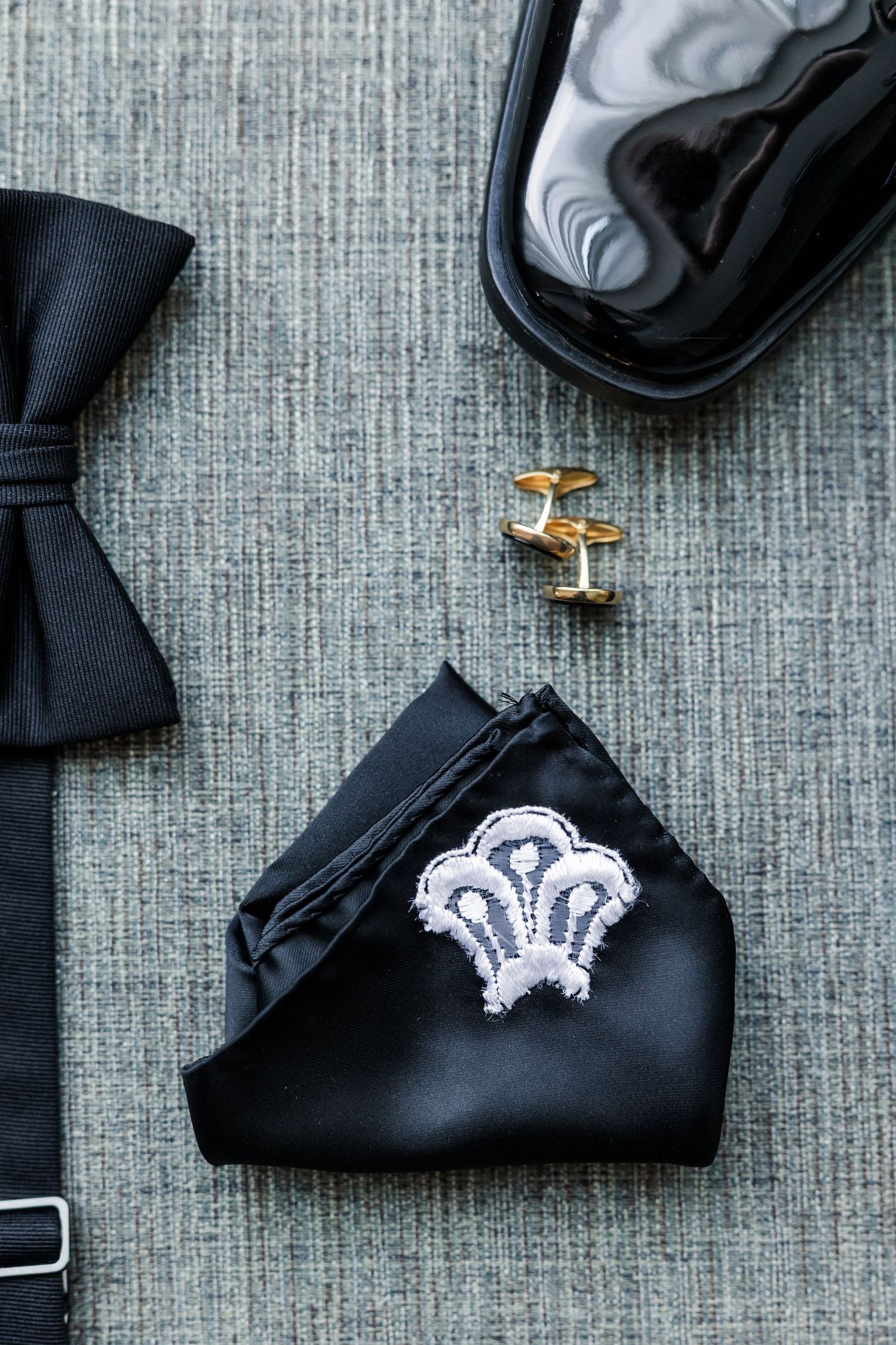 Black bow tie, gold cufflinks, embroidered pocket square, and patent leather shoes arranged on gray fabric.
