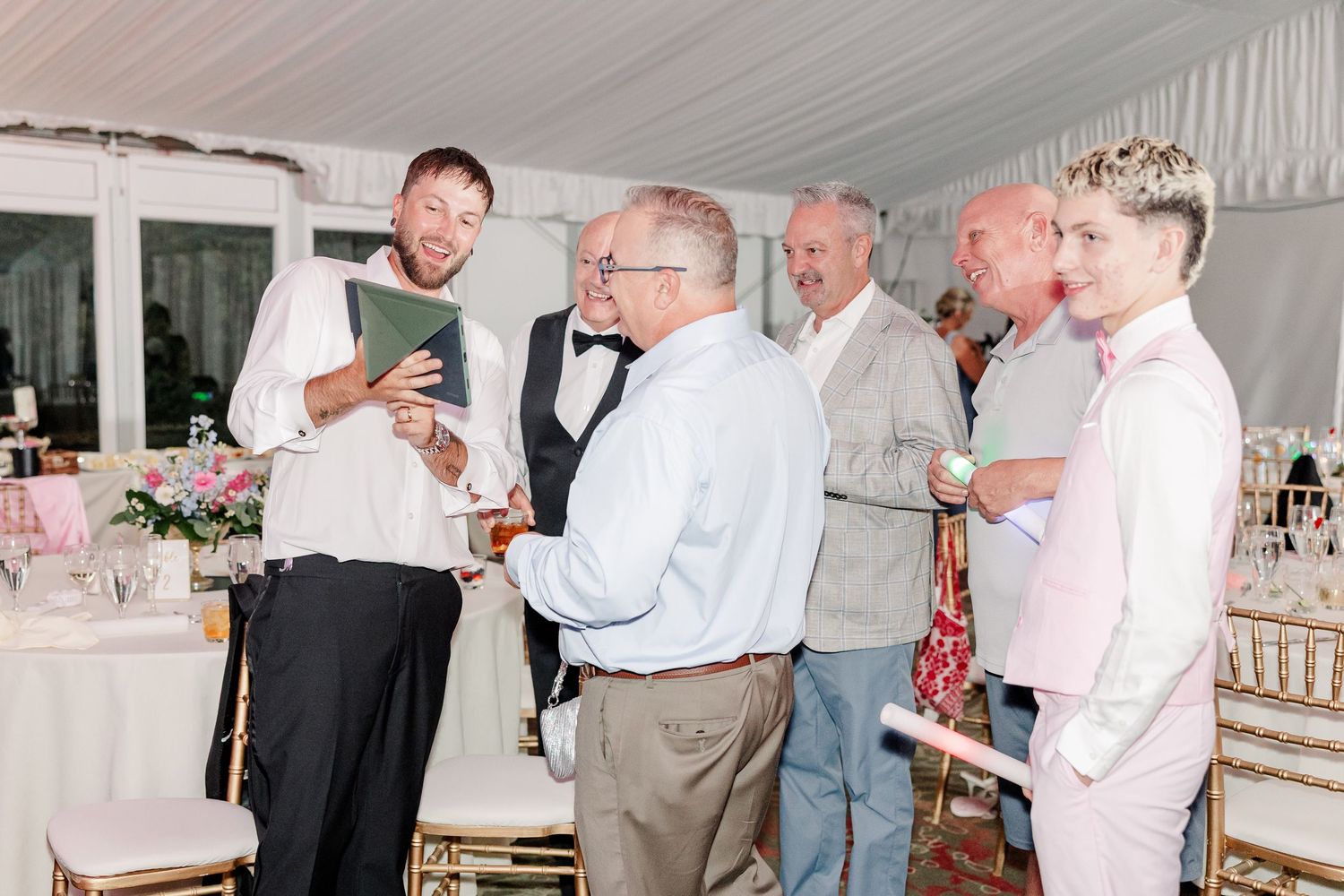 Group of male wedding guests gather and converse while reviewing something on a tablet at reception.