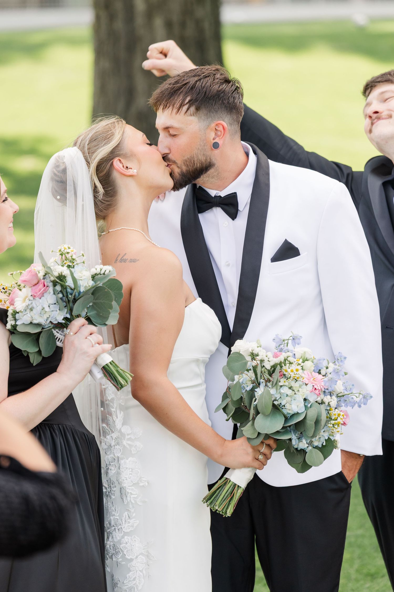 Bride and groom sharing first kiss while wedding party celebrates around them outdoors.