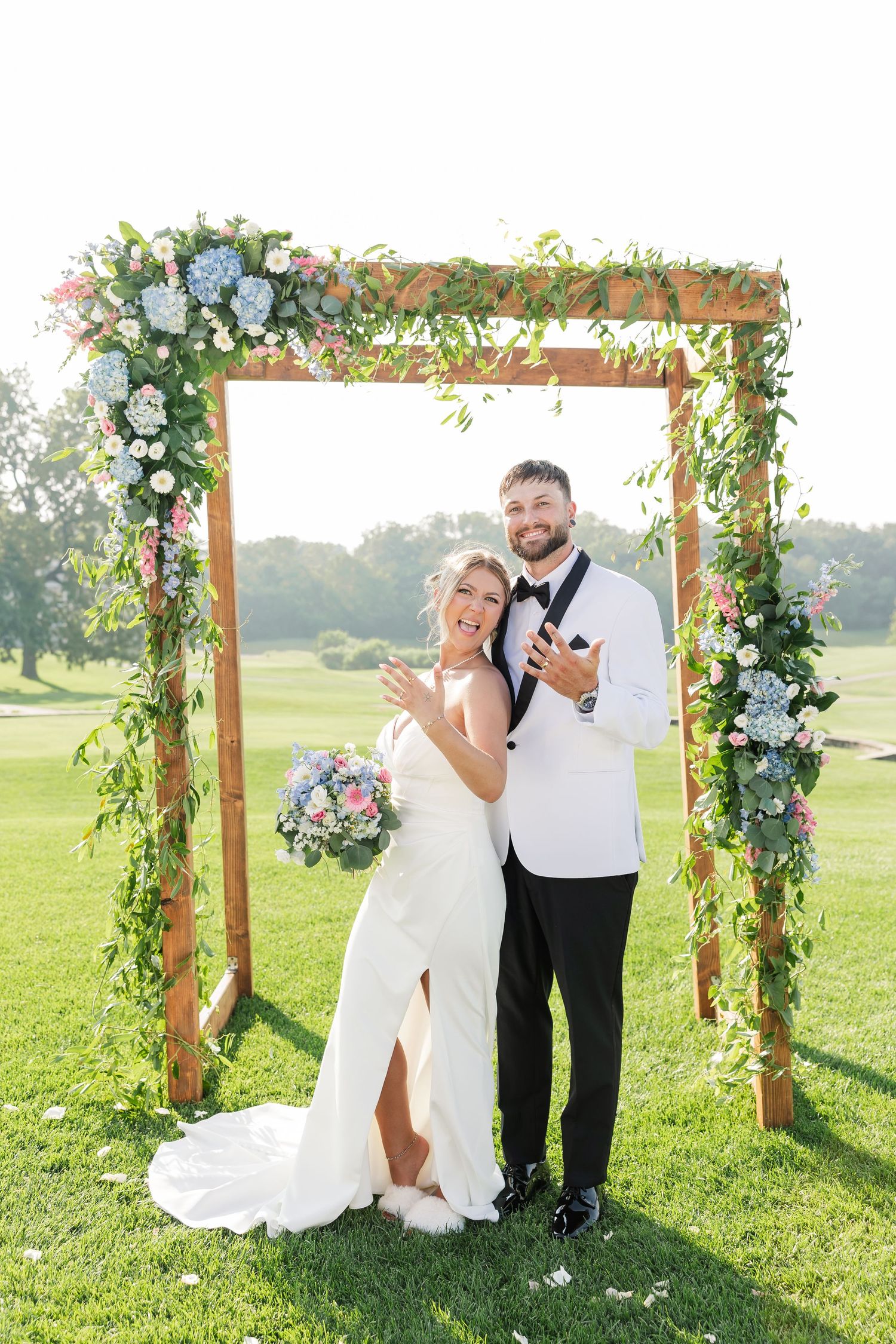 Smiling bride and groom pose under a wooden floral arch adorned with hydrangeas and greenery on a golf course.