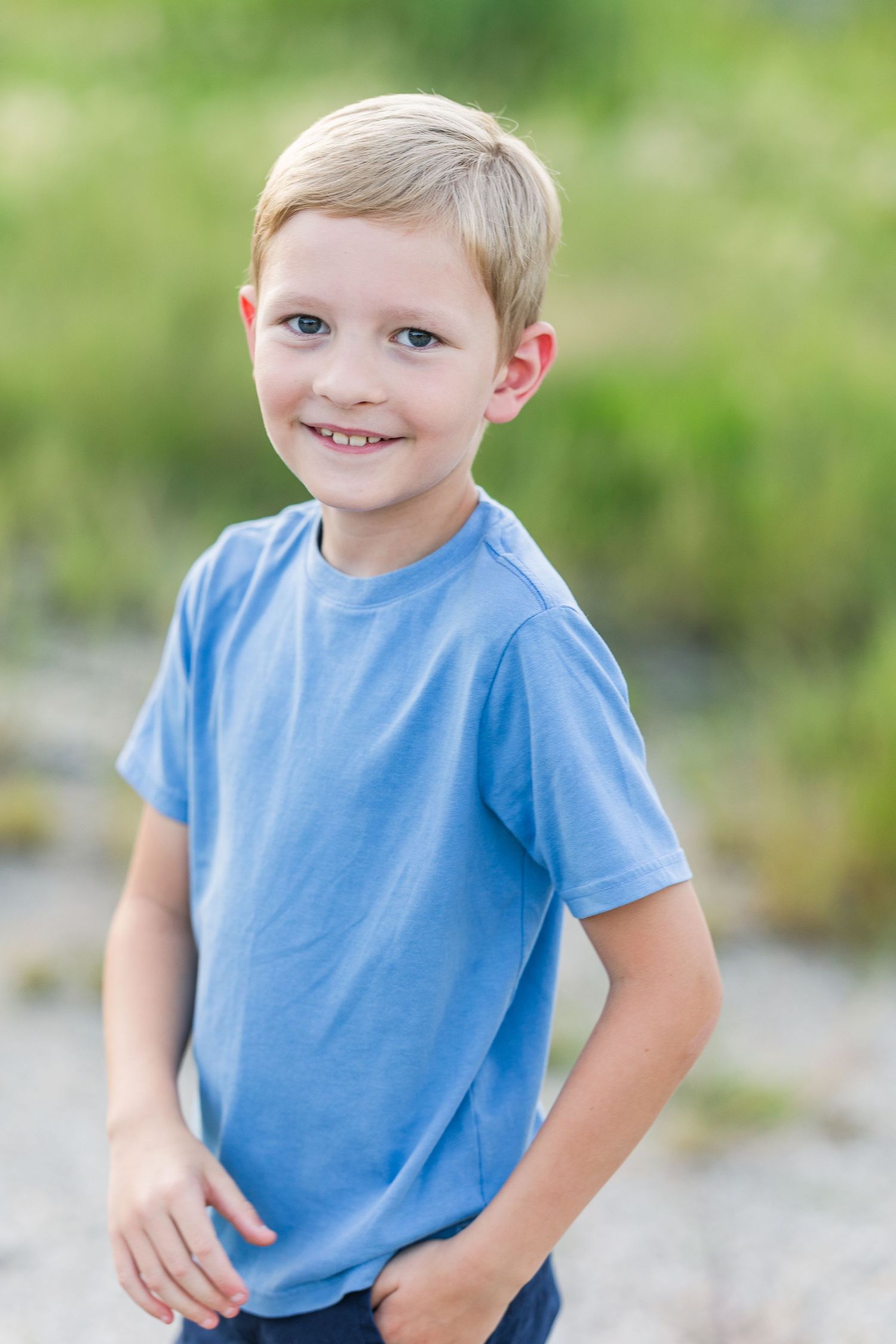 A young child in a light blue t-shirt poses outdoors with a playful expression against a blurred natural background.