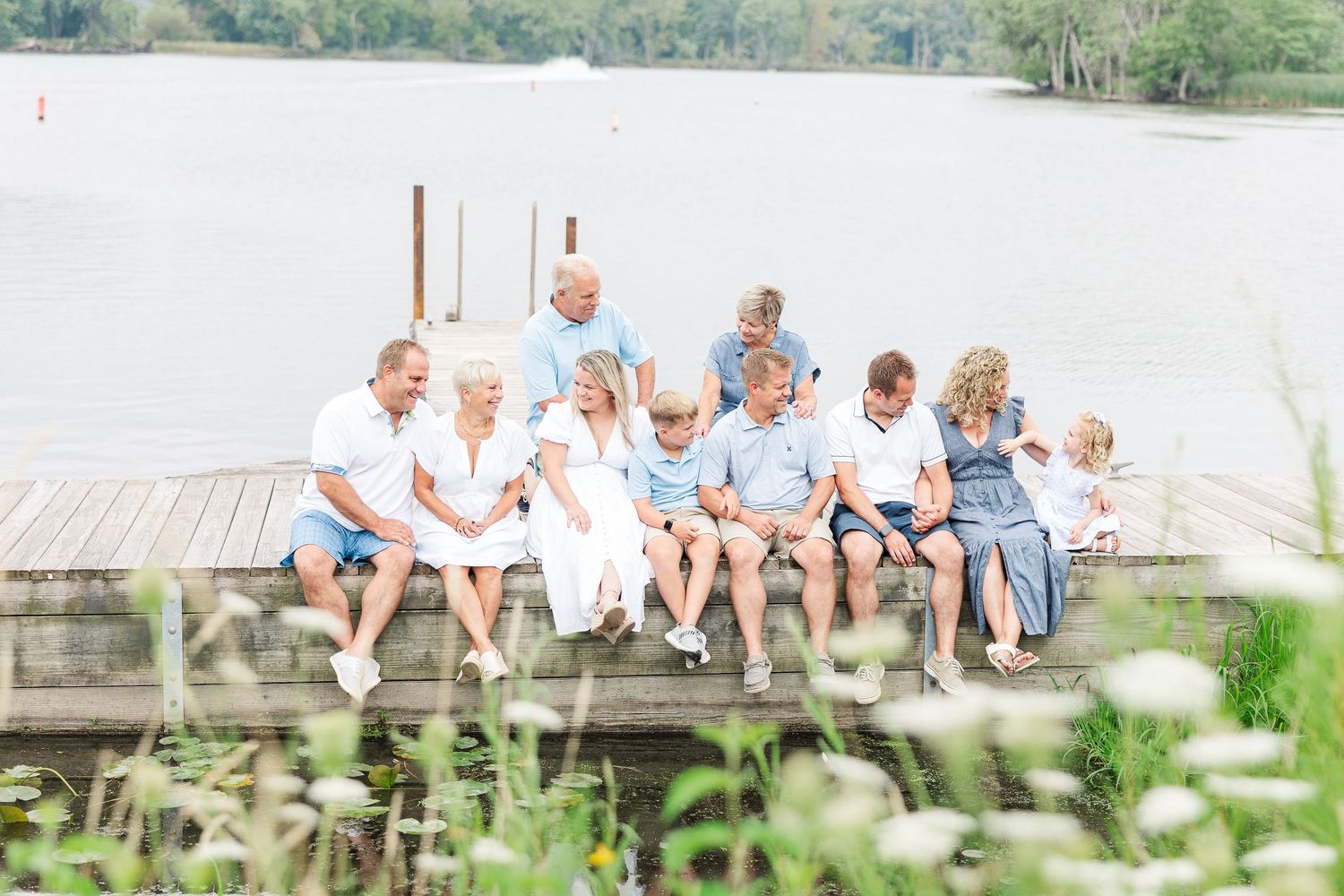 Extended family gathering on wooden pier overlooking peaceful lake with natural vegetation in foreground.