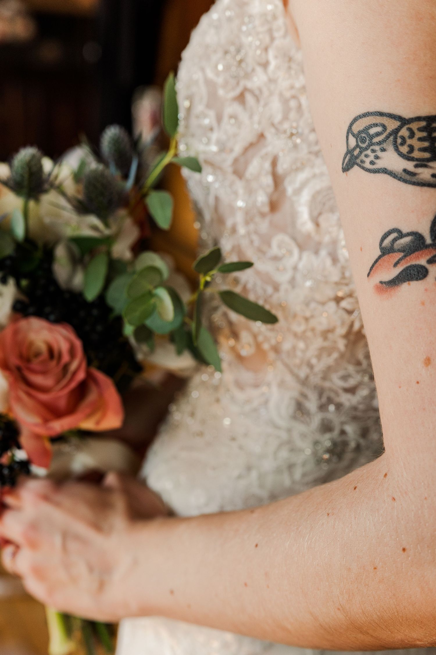 Close-up of a bird tattoo on an arm holding flowers with white lace in the background.