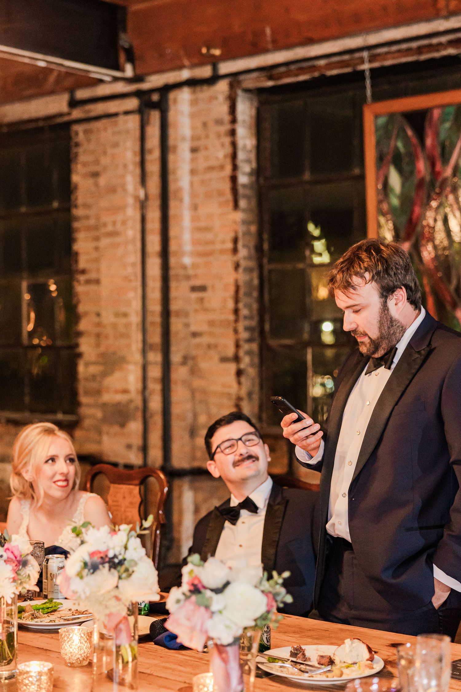 Wedding reception guests sharing moment at rustic wooden table with floral centerpieces.
