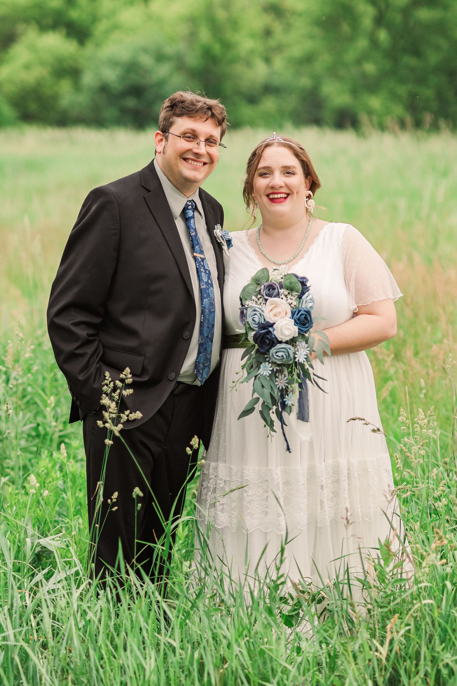 Newlyweds pose together in a lush green field with bride holding blue and white bouquet.