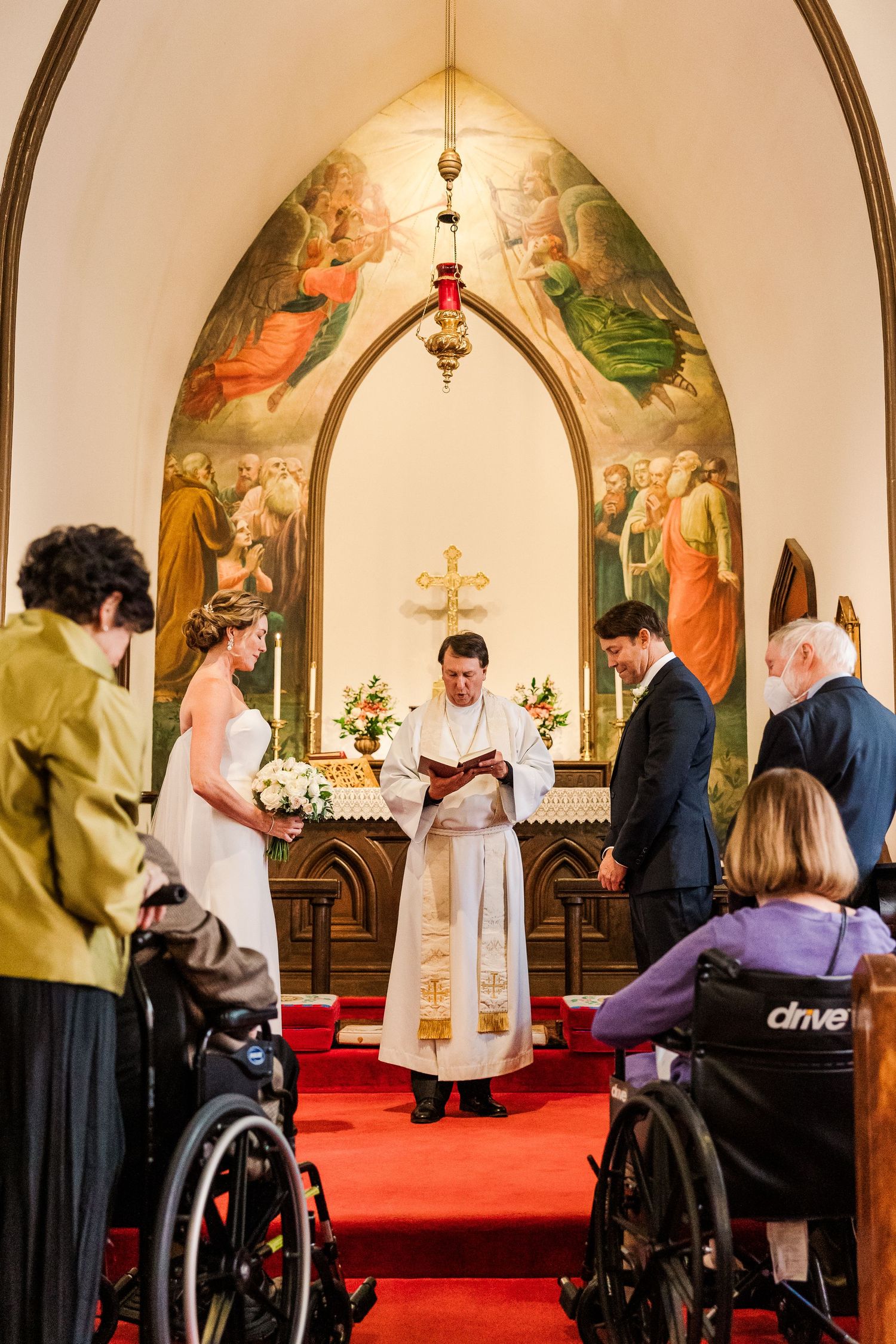 A wedding ceremony takes place in a historic church with wheelchair guests and religious murals on the walls.
