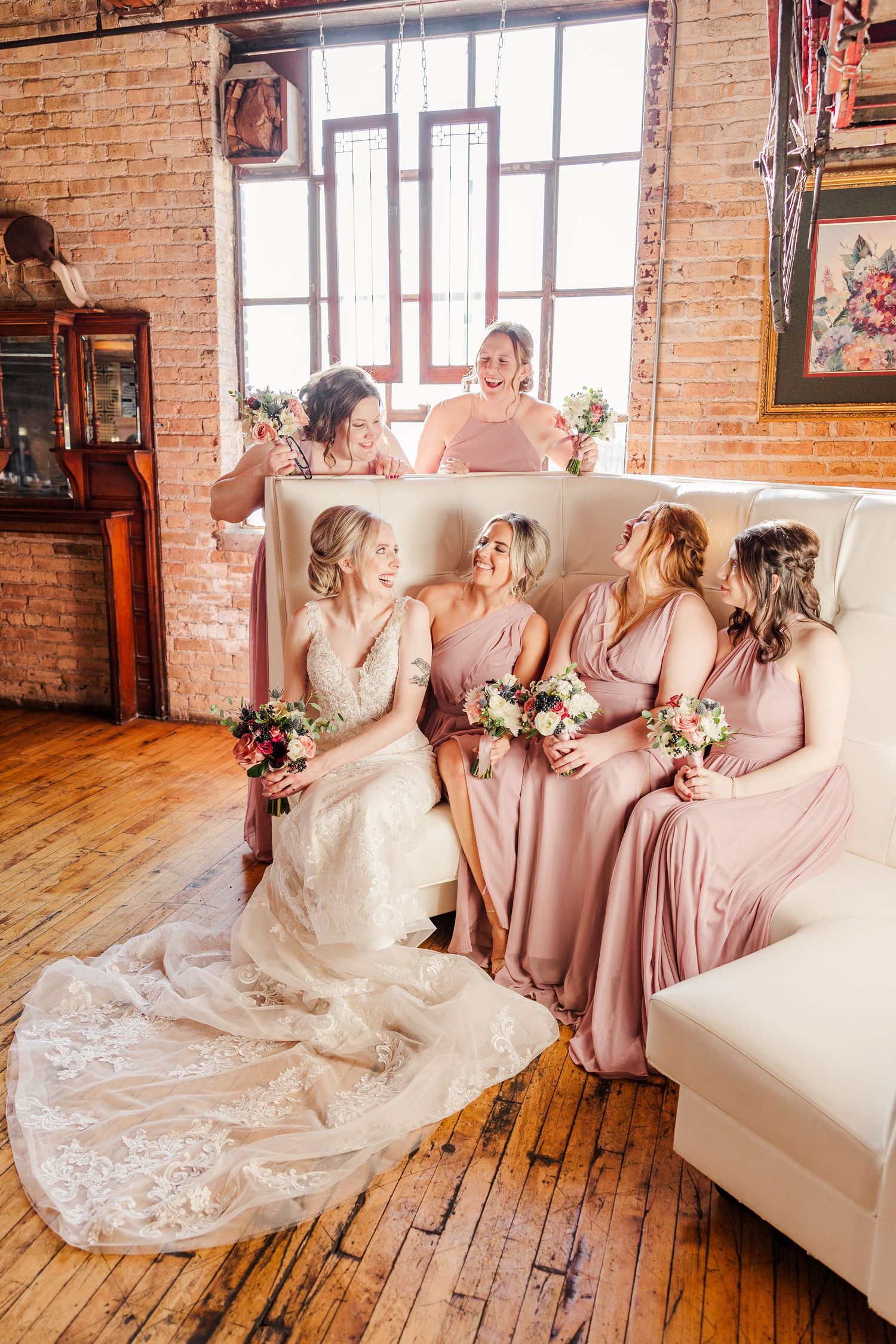 Wedding party enjoying candid moment on white sectional with exposed brick backdrop.