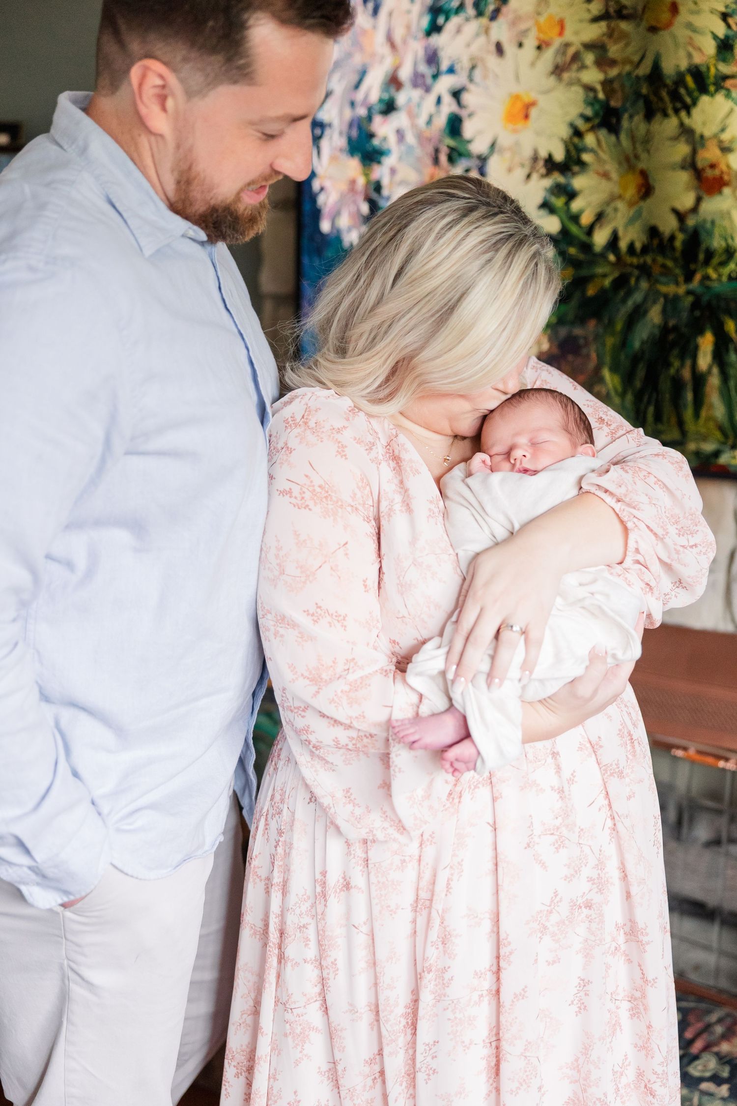New parents tenderly hold their sleeping newborn baby, mother in floral pink dress with father in light blue shirt.