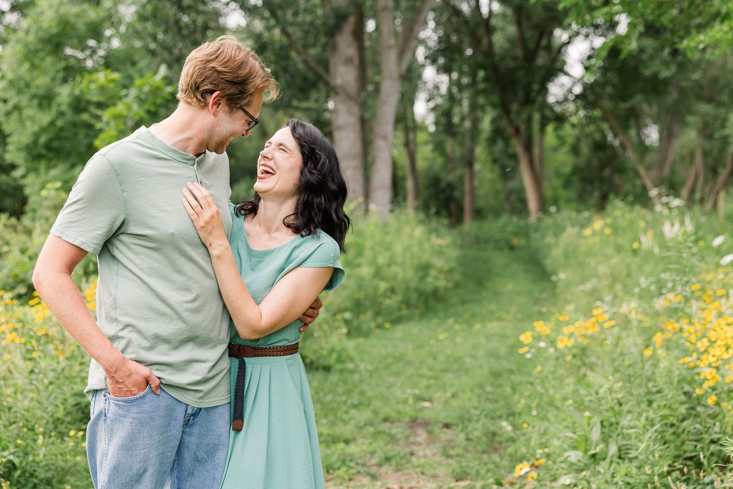 A laughing couple embraces in a lush green meadow with yellow wildflowers during an outdoor engagement session.