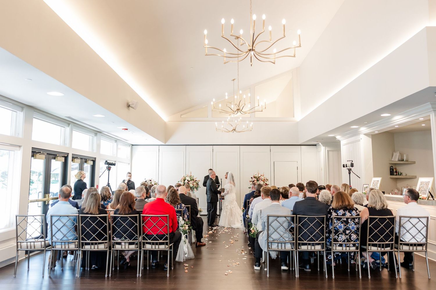 Wedding guests seated in rows watch a bride and groom at the altar in a bright ceremony room with chandeliers and large windows.