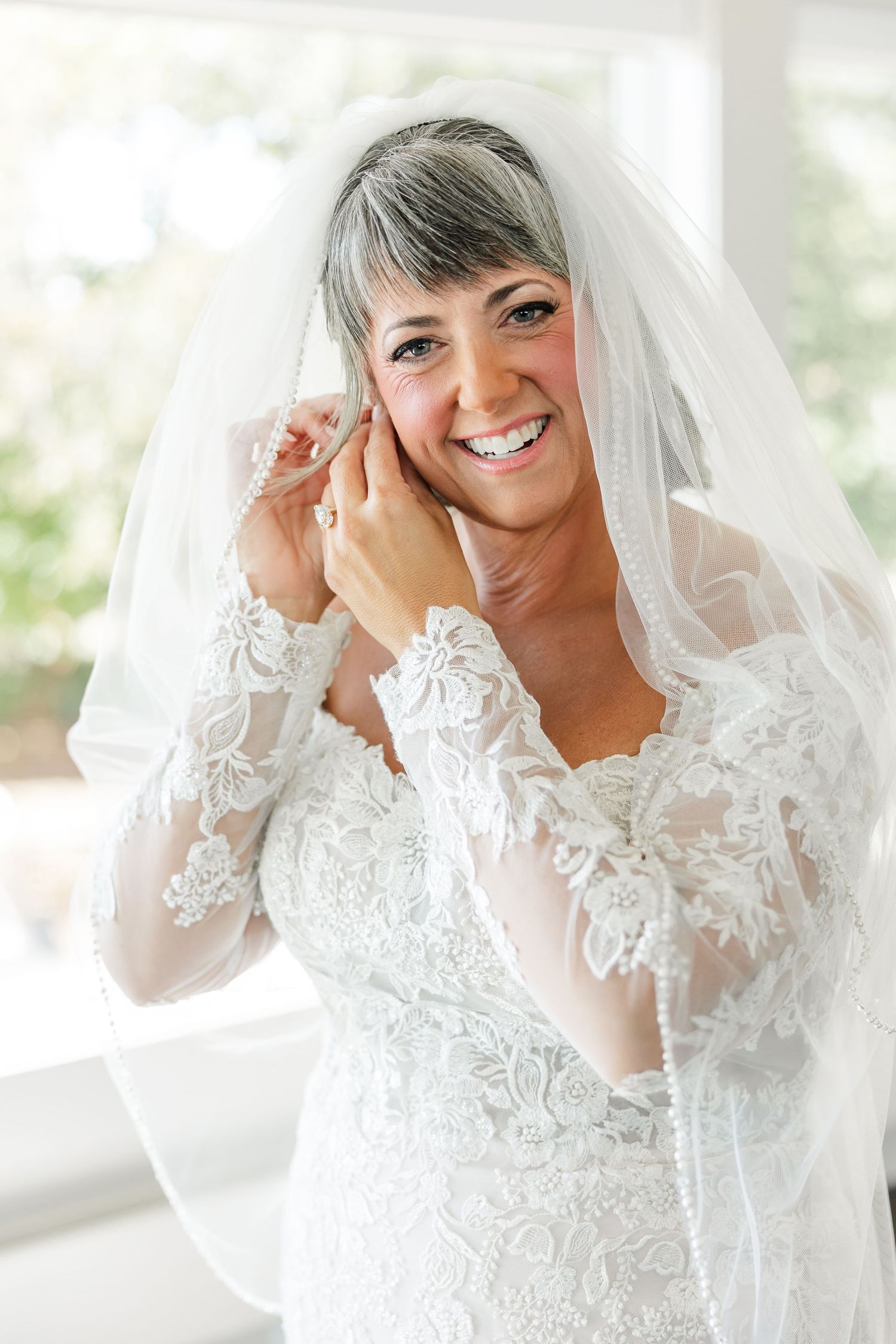 Smiling bride in a long-sleeve lace wedding gown and sheer veil adjusts her earring in front of a bright window.