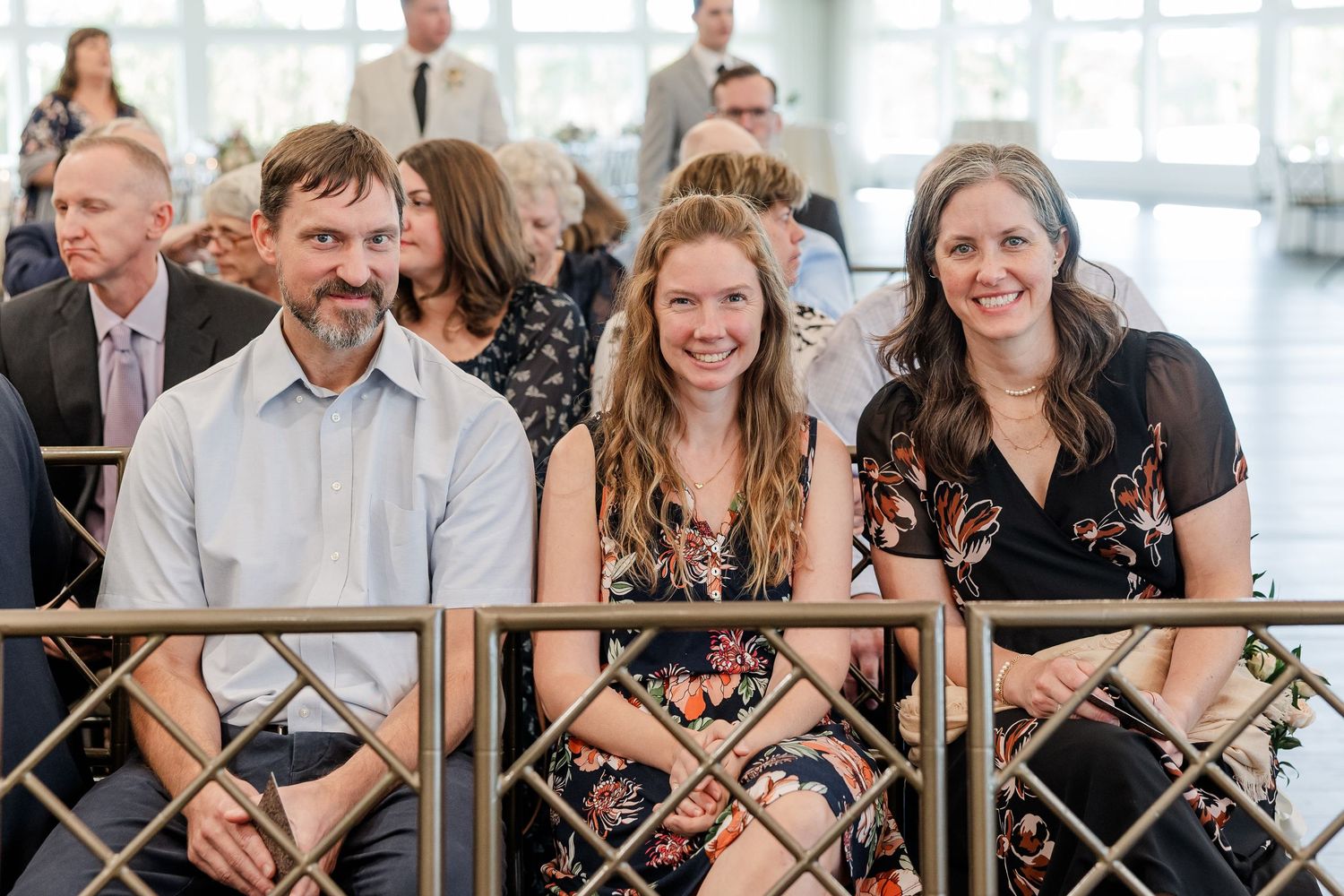 Three smiling adults sit side by side in a bright hall behind a decorative metal railing, with other guests gathered in the background.