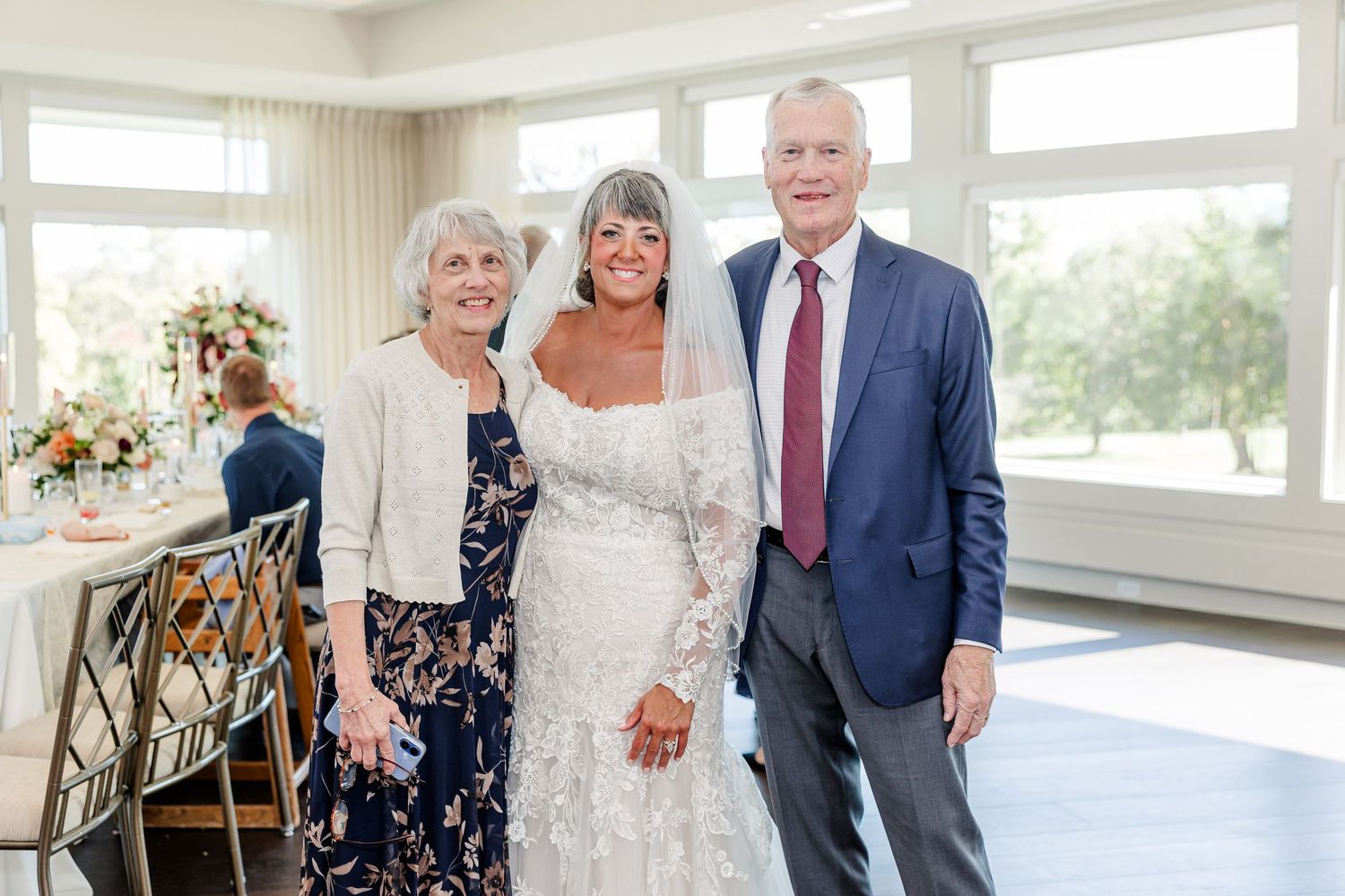 Bride in a lace wedding gown and veil poses with an older woman and a man in a blue suit at a bright wedding reception venue.