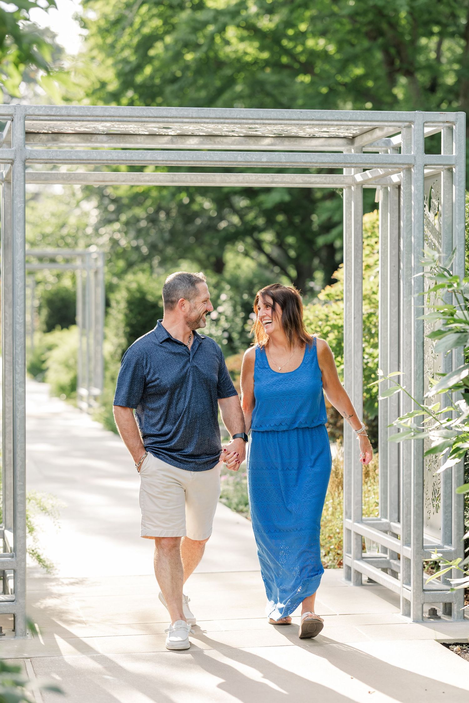 Smiling couple walks hand in hand under garden pergola on sunny day.