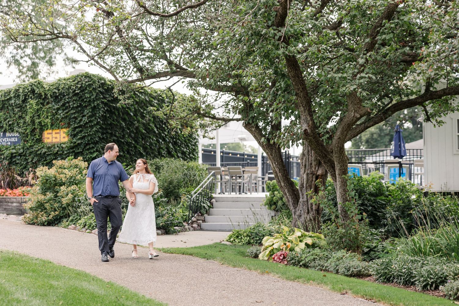 Couple walking hand in hand through a lush garden path surrounded by green trees and colorful flower beds.