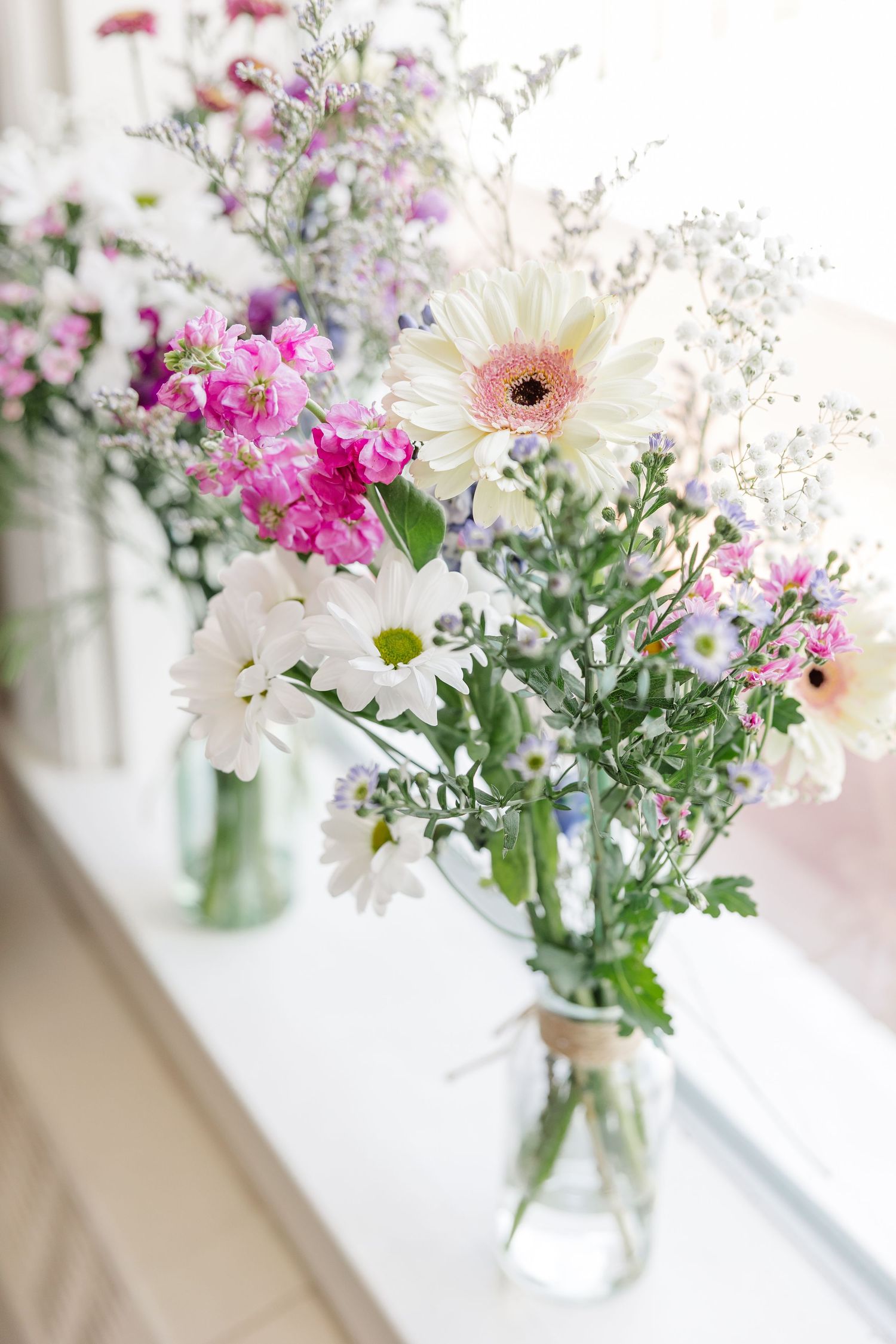 Romantic arrangement of white gerbera daisies, pink flowers, and wildflowers in glass vases on a windowsill.