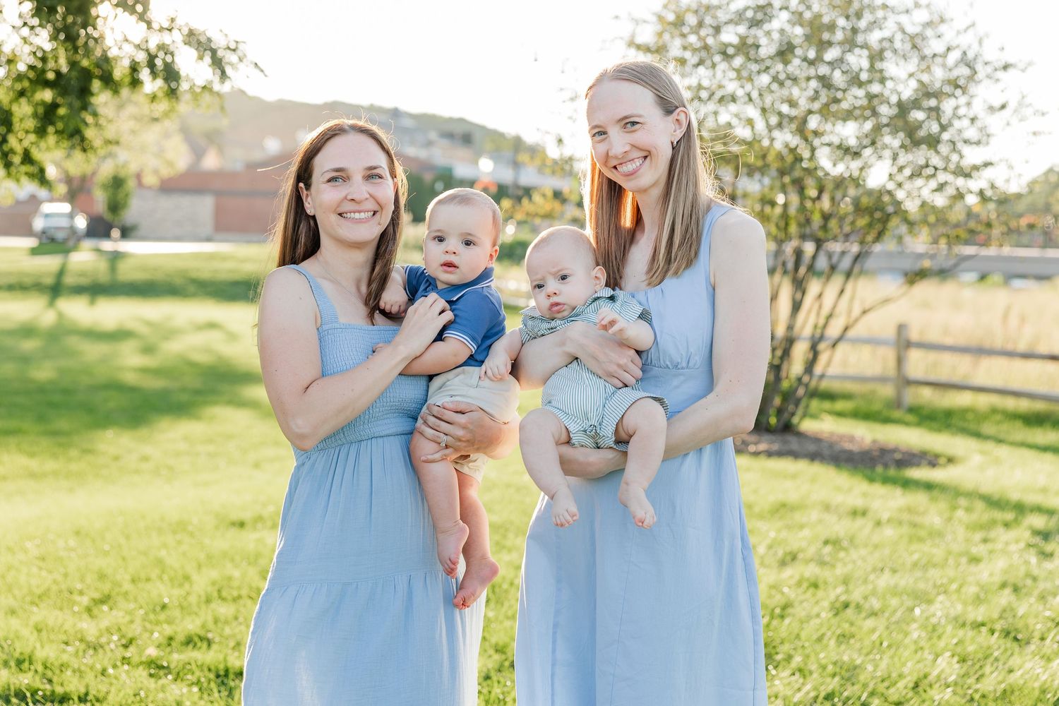 Two women in light blue dresses holding babies and smiling together on a sunny green lawn outdoors.
