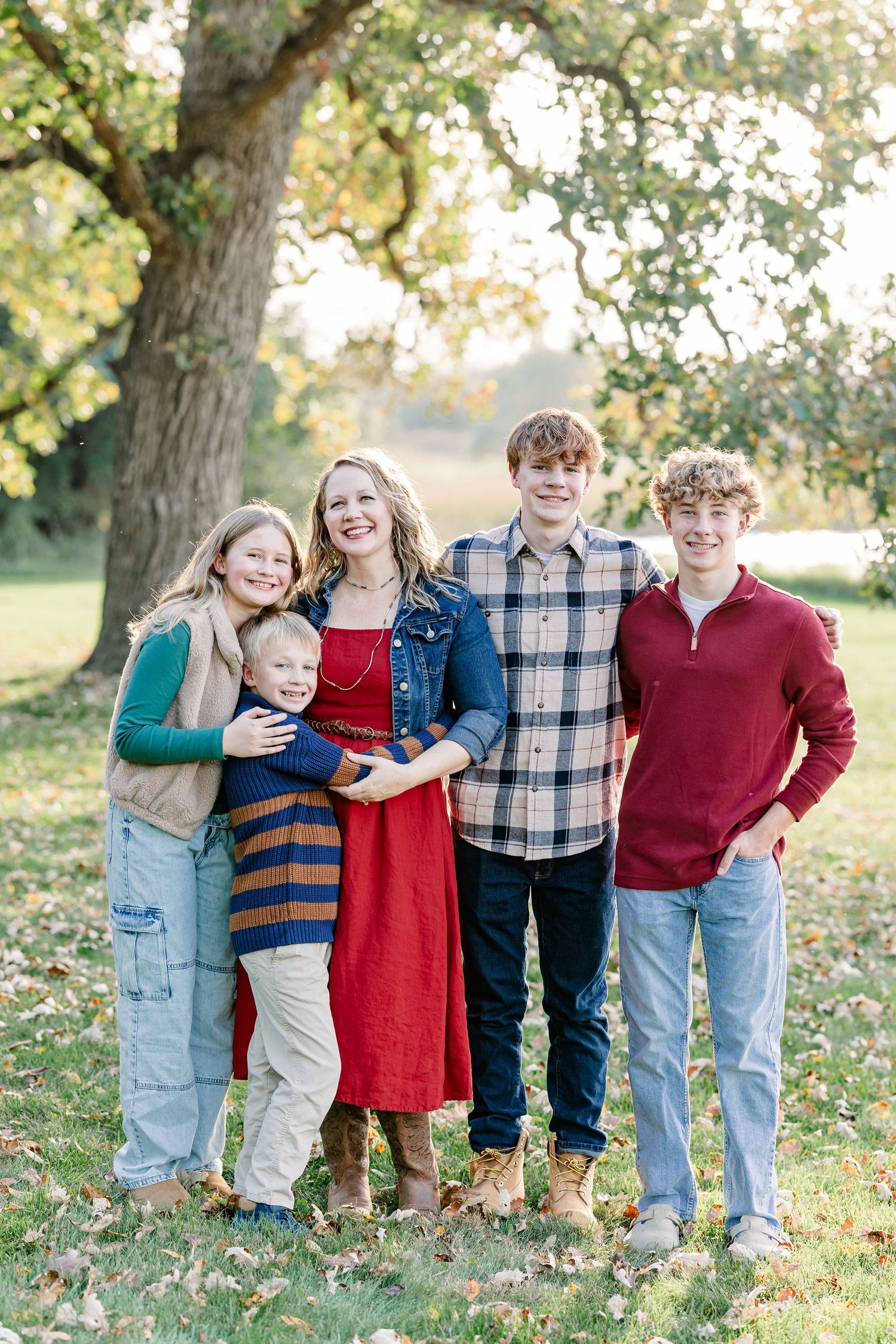 Mother in a red dress and denim jacket stands with her four smiling children under a large tree on a leaf-covered lawn.