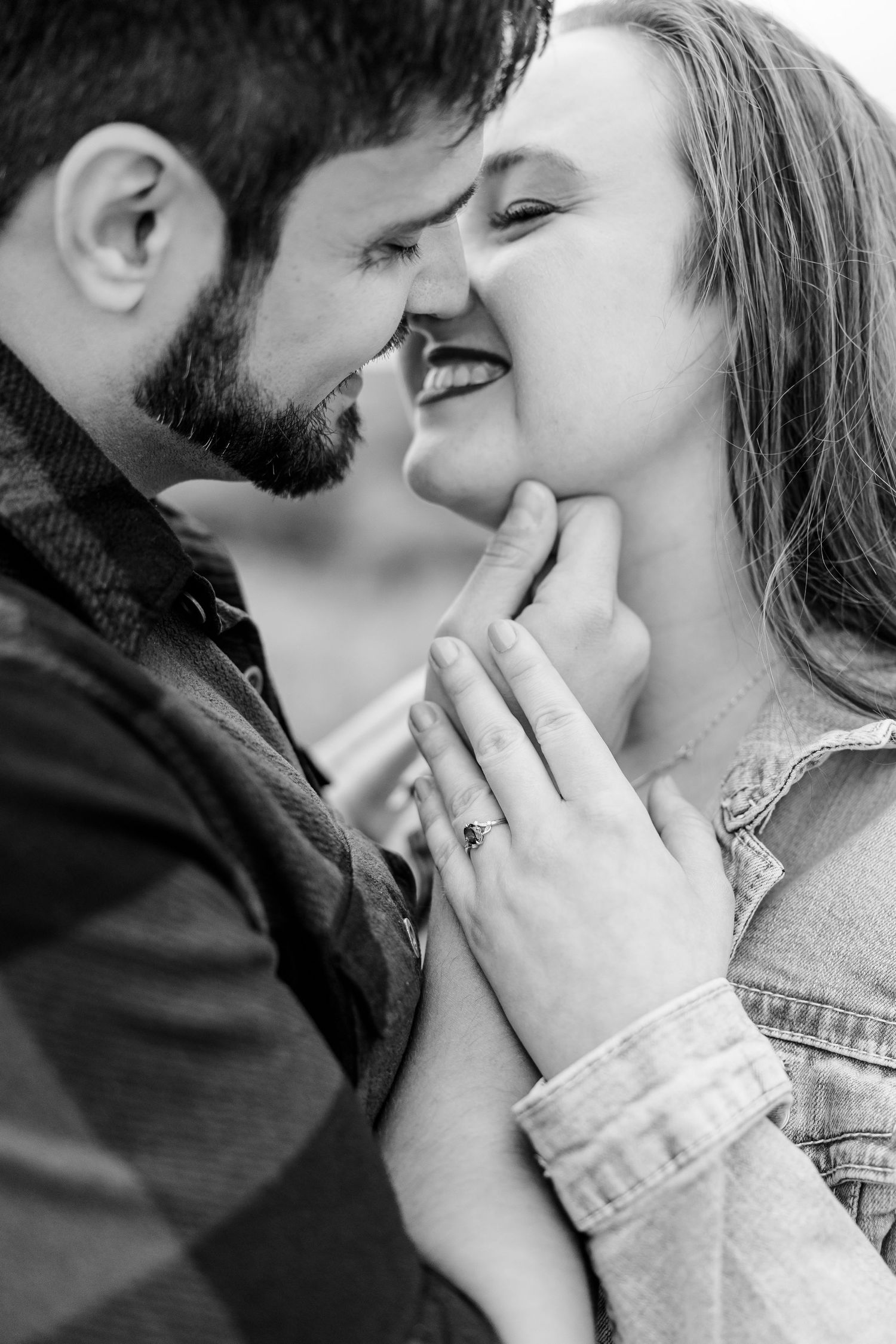 Black and white close-up of a laughing couple nose to nose, her hand on his chest displaying an engagement ring.