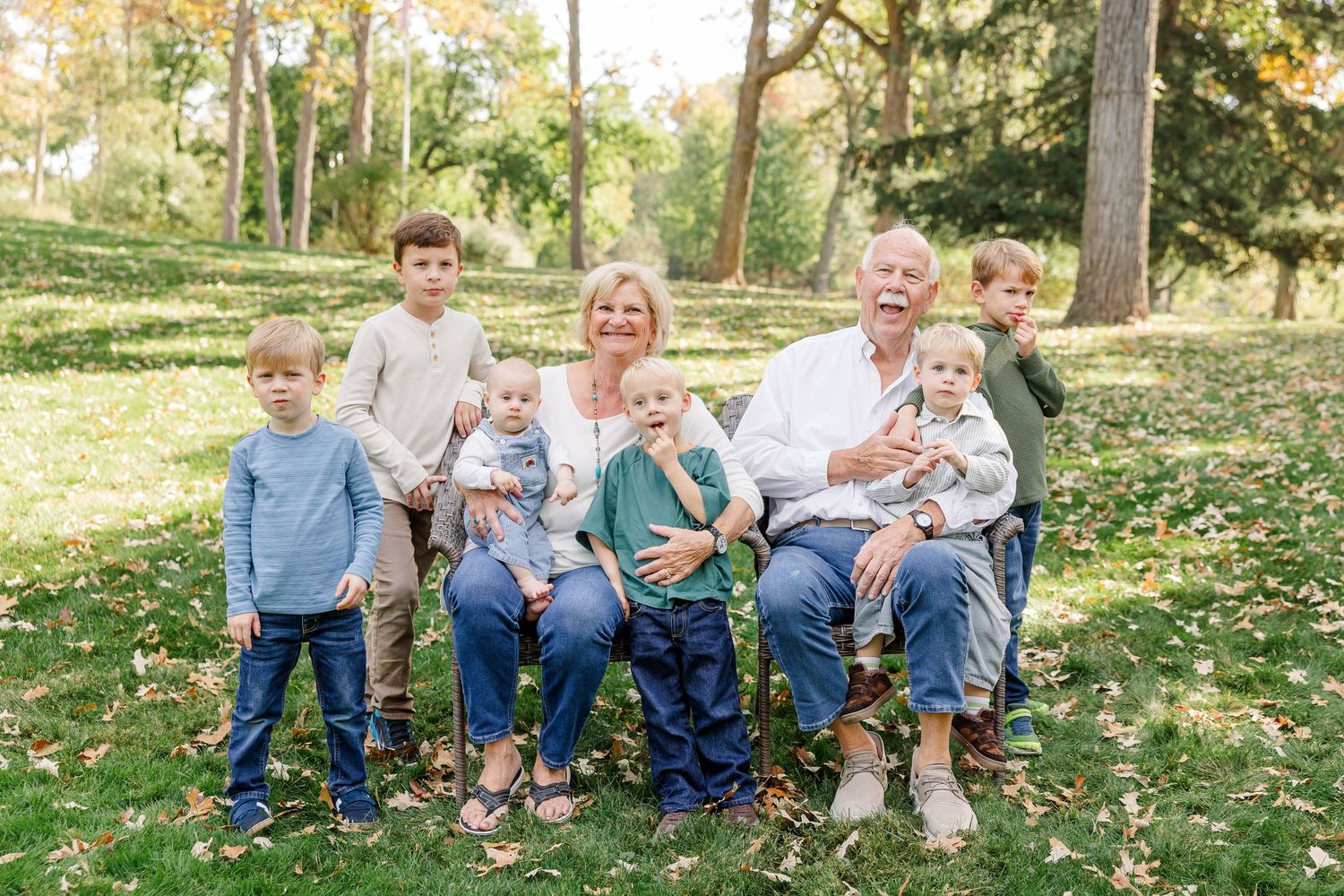 Grandparents sit outdoors in a park with six young grandchildren gathered around them on a sunny fall day.