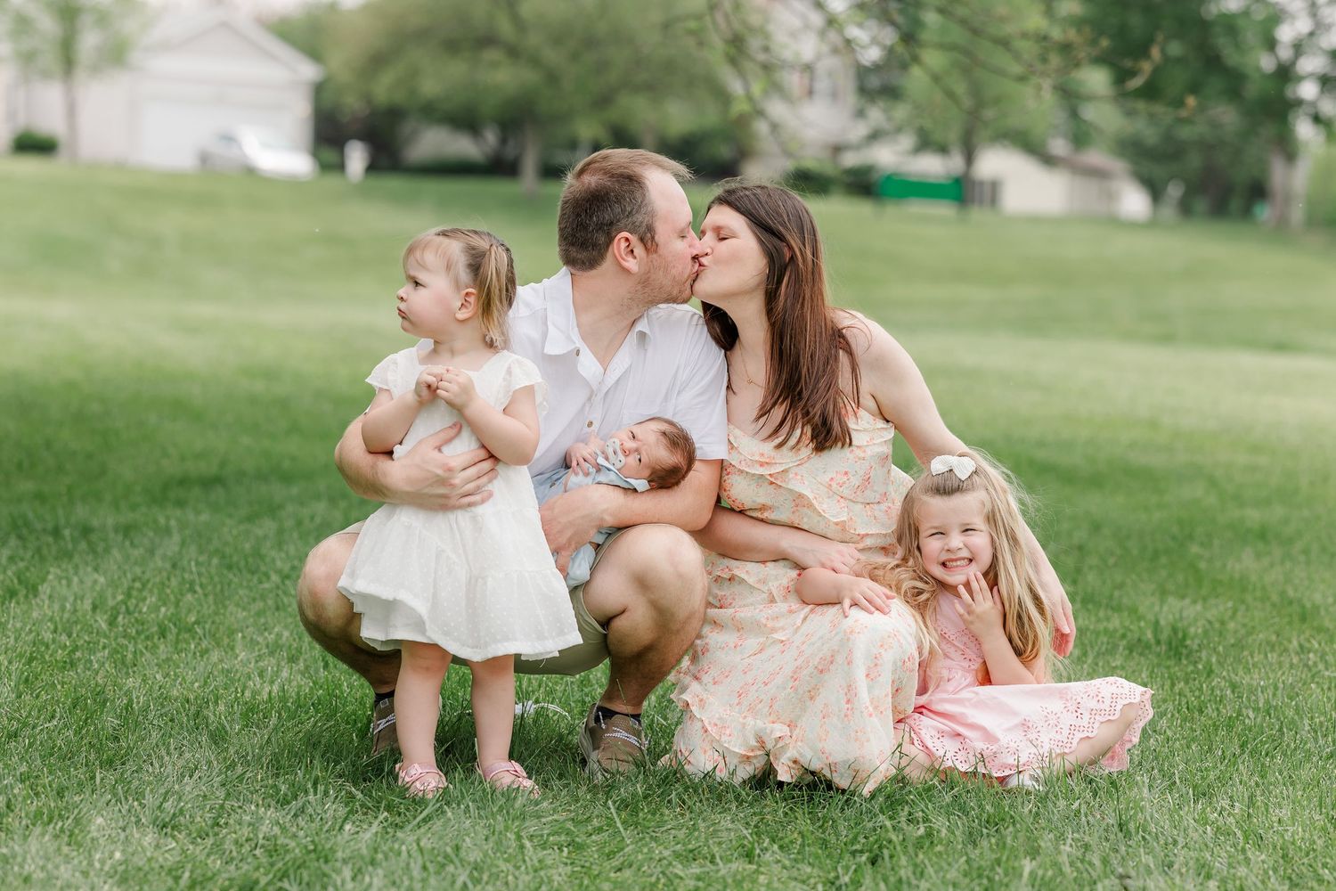A couple shares a kiss while crouching on green grass surrounded by their three young children during a family photo session.