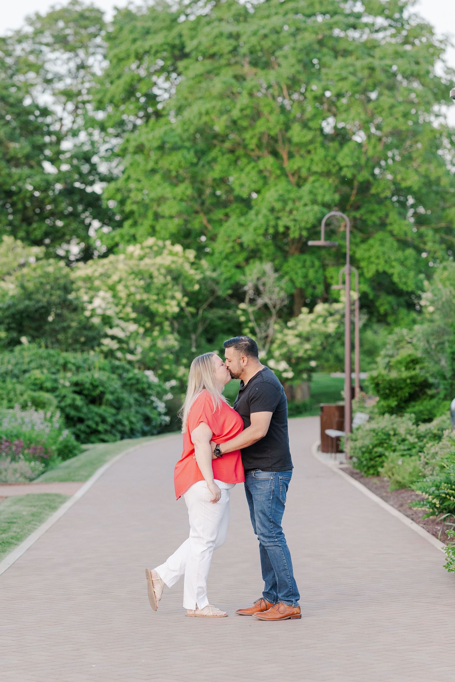 Romantic embrace on garden path with green trees and lamp post in background.