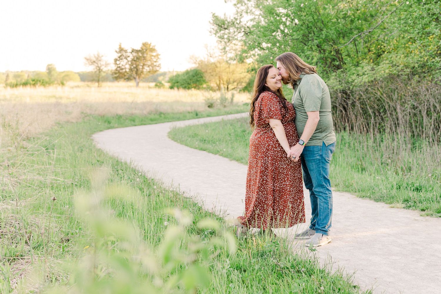 A couple embraces on a winding dirt path surrounded by green grass and golden sunlight.