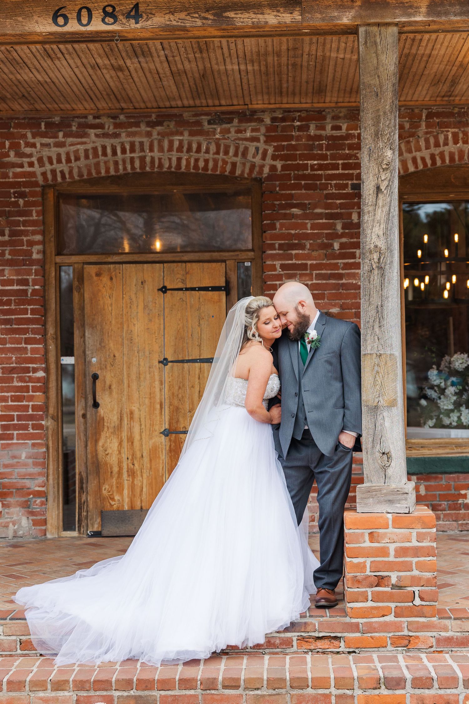 Romantic wedding portrait against vintage brick building with wooden door and string lights.