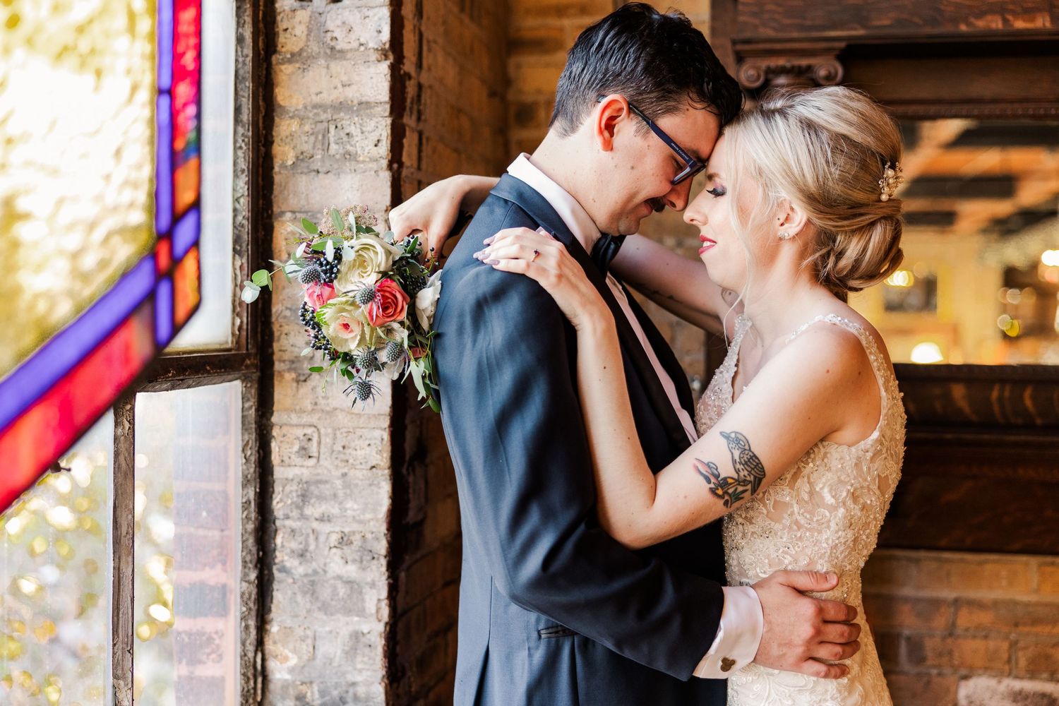A wedding couple shares an intimate moment near a stained glass window in a rustic brick venue.