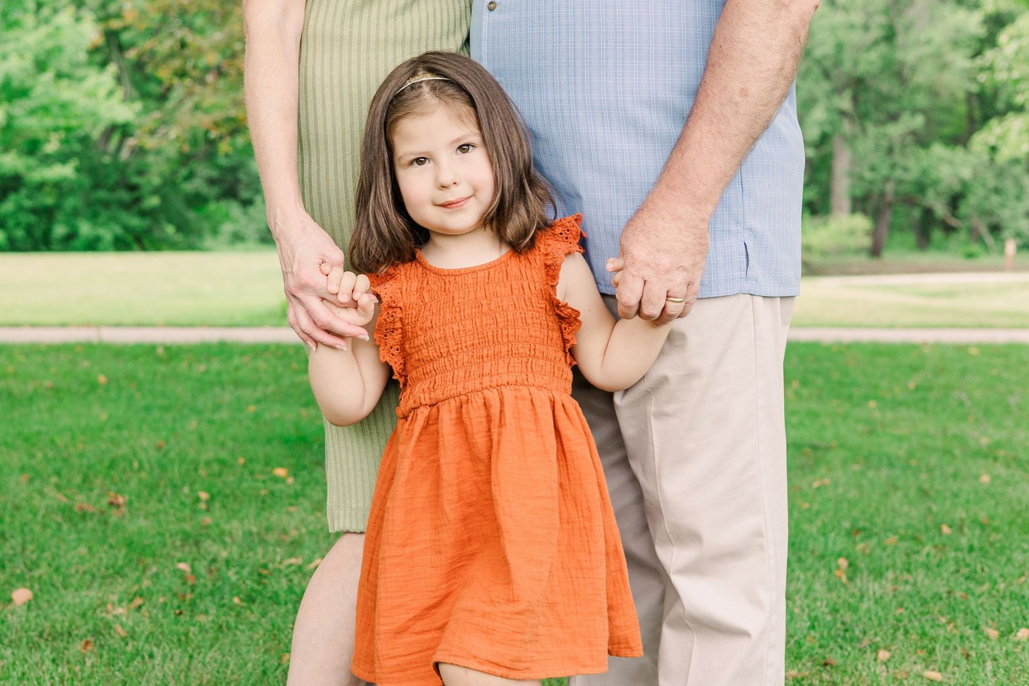 A small child in an orange dress holds hands with adults while standing in a grassy park.