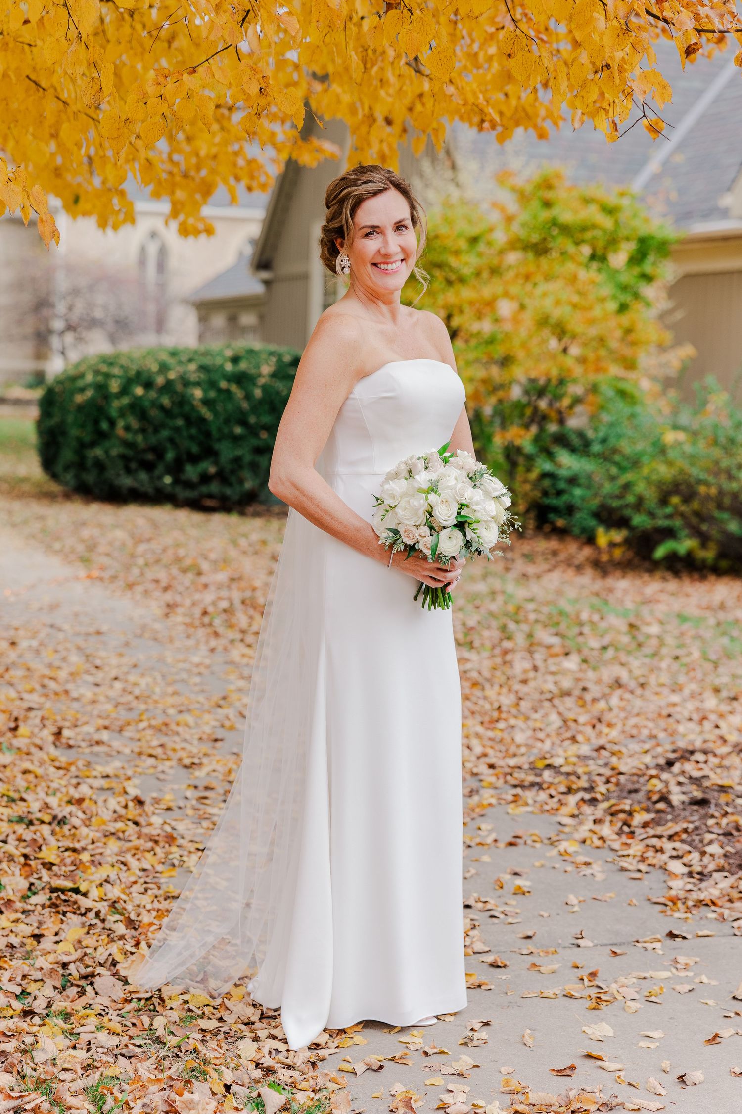 Elegant bridal portrait in a strapless white wedding dress holding a white bouquet amid golden autumn leaves.