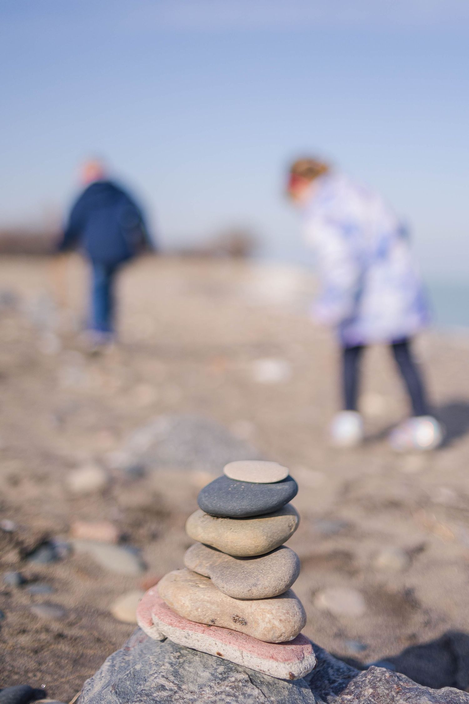Stone cairn in foreground with blurred figures walking on rocky beach.