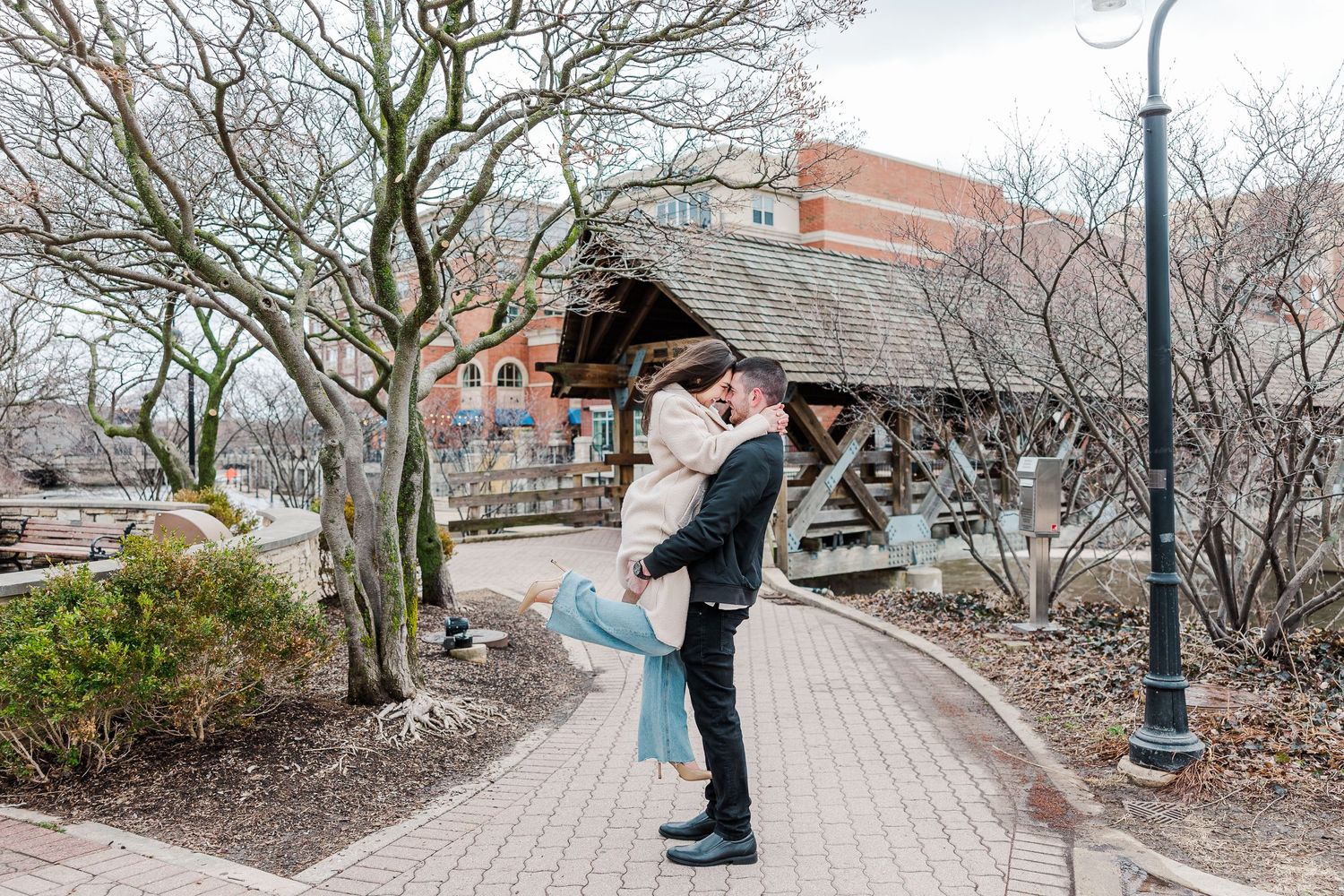 A romantic lift pose on a curved brick path with bare winter trees and a wooden shelter.