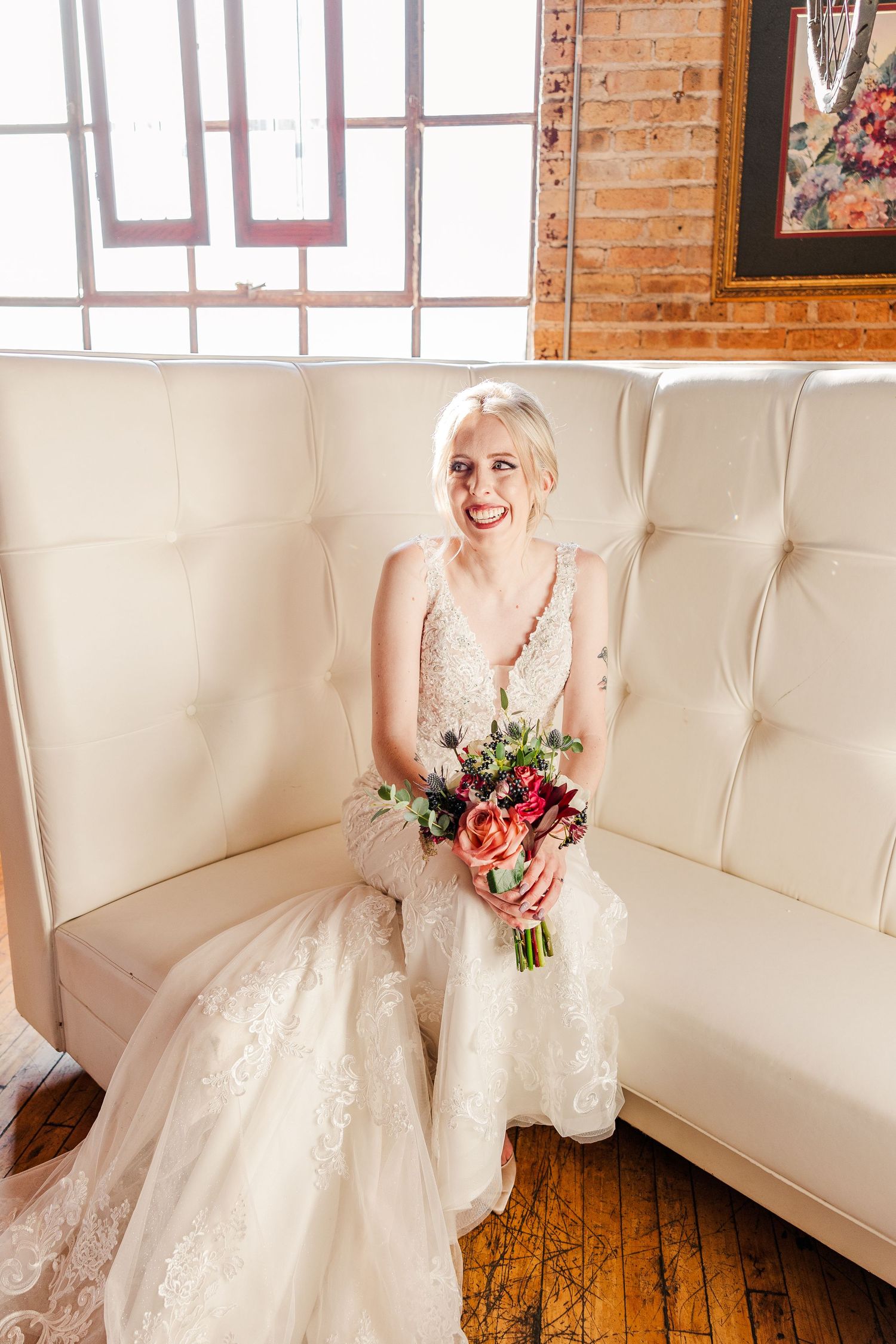 Elegant bridal portrait in lace gown with floral bouquet against exposed brick wall.